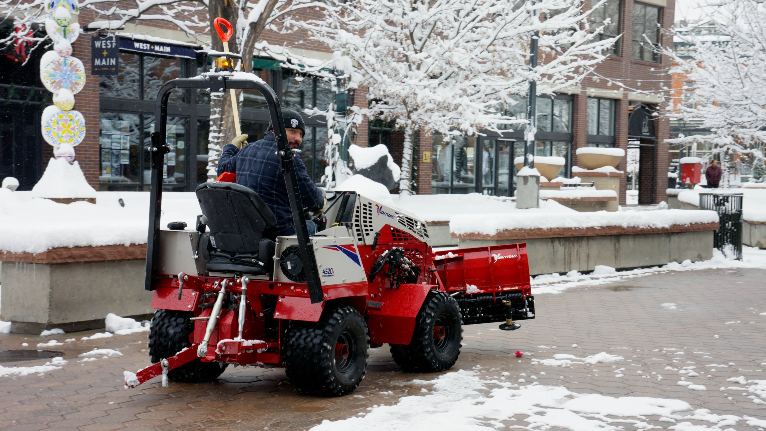 A man operating a small red snow removal machine on a city sidewalk, with snow-covered trees and buildings in the background.