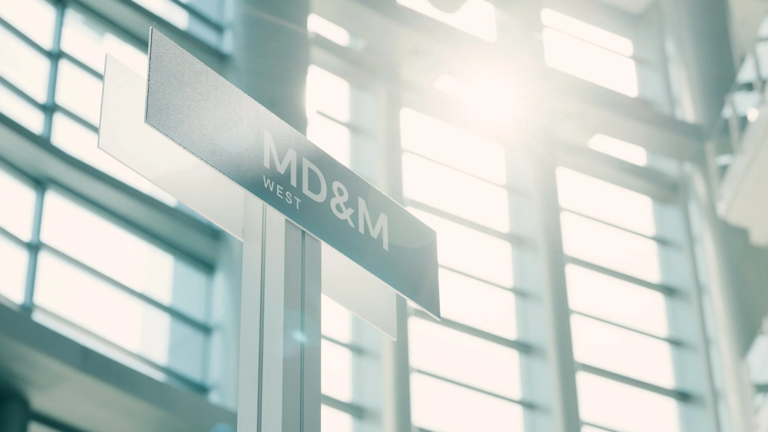 A glass door sign at an airport gate with the text 'MD&M WEST' visible. The background shows large windows allowing sunlight to stream in, creating a bright and airy atmosphere.