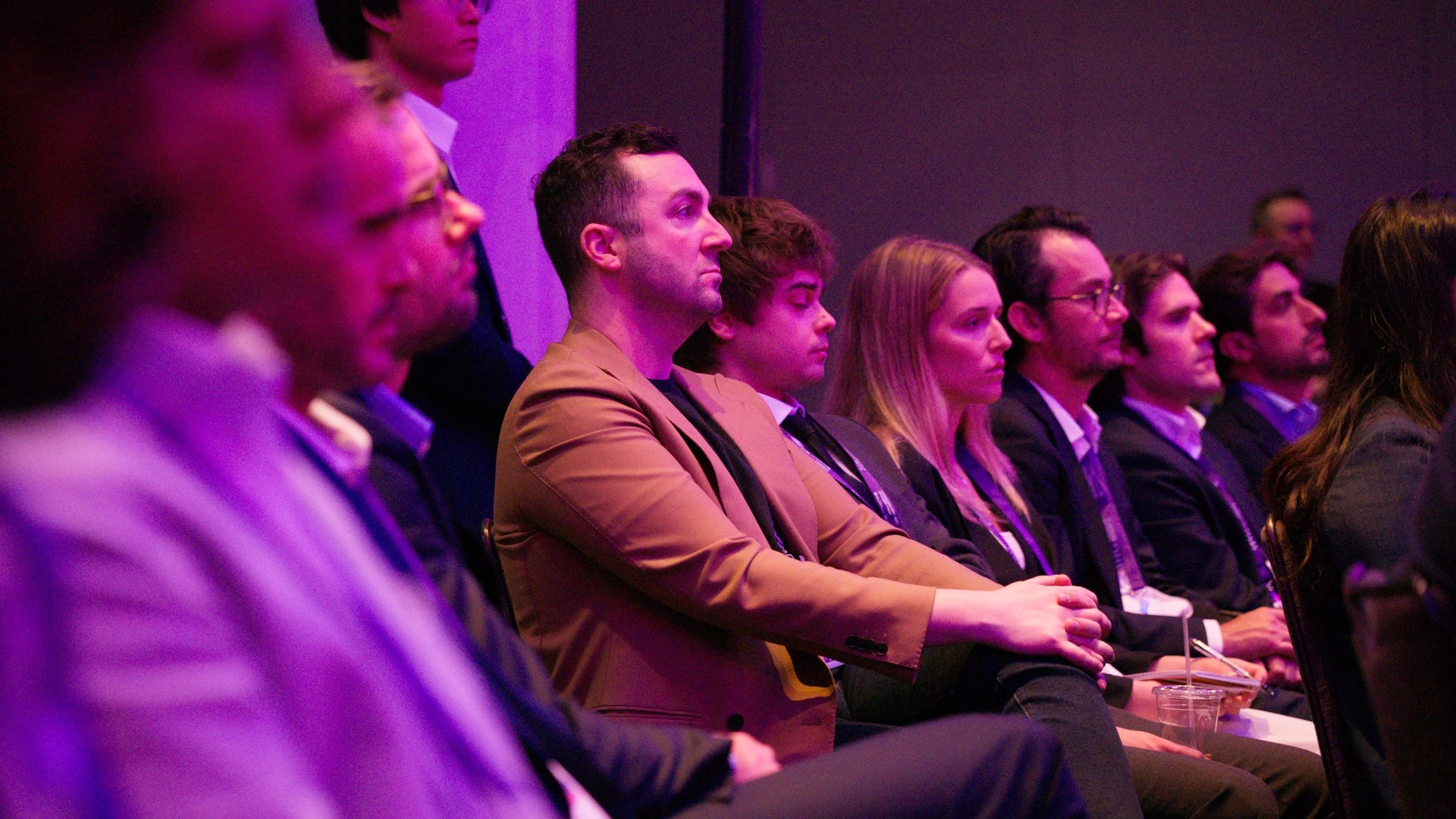 People sitting in an audience at an event, with some individuals focused and attentive, illuminated by purple lighting.