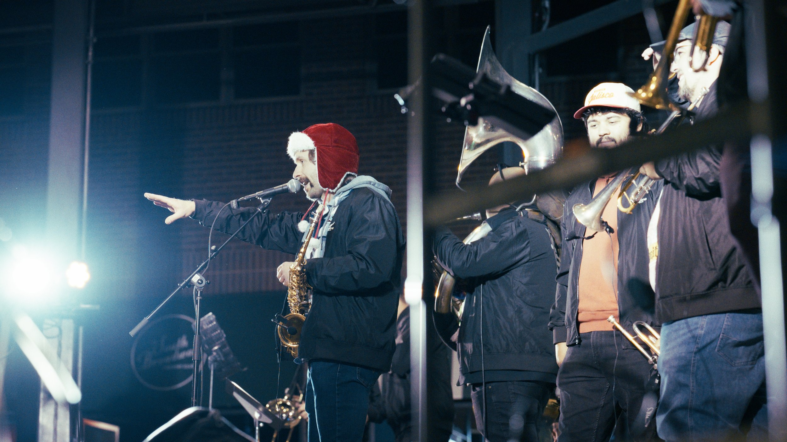 Musicians on stage, including a man in a Santa hat speaking into a microphone, holding a saxophone, with others playing brass instruments, during a holiday-themed performance.