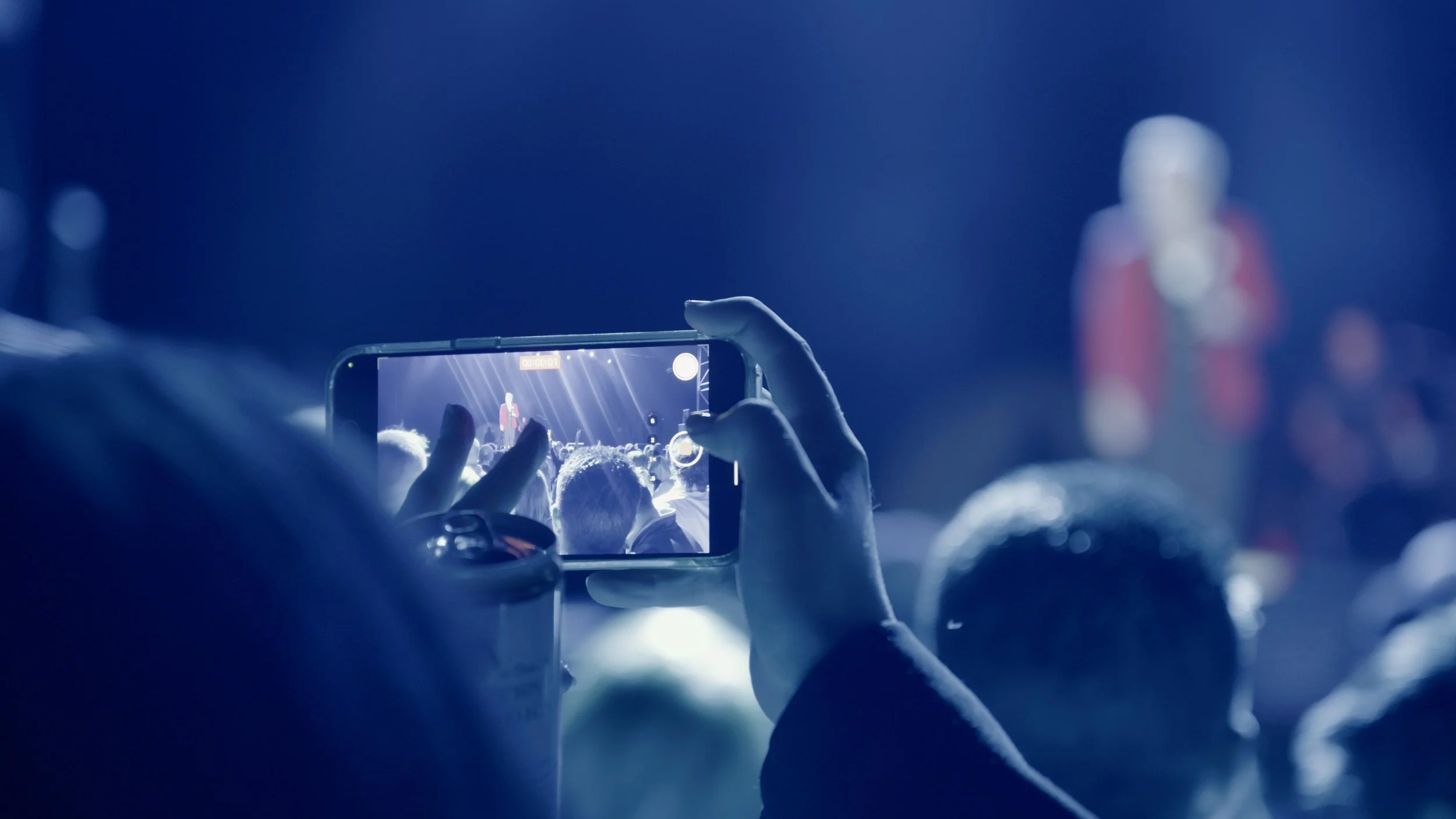 A person holding up a smartphone, recording a live performance on stage with bright lights, while the audience watches. The background is blurred with a focus on the phone screen showing the performer in red.