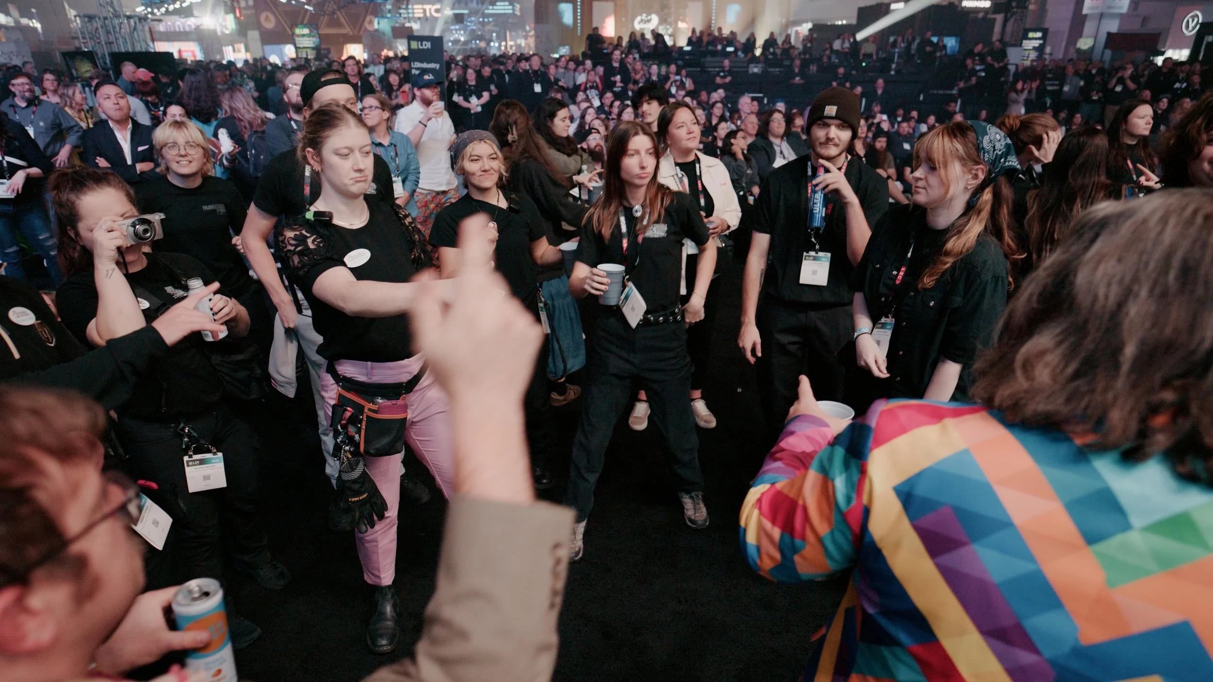 A large crowd of people gathered at an indoor event, some taking photos and wearing event badges. The background features various signs and banners.