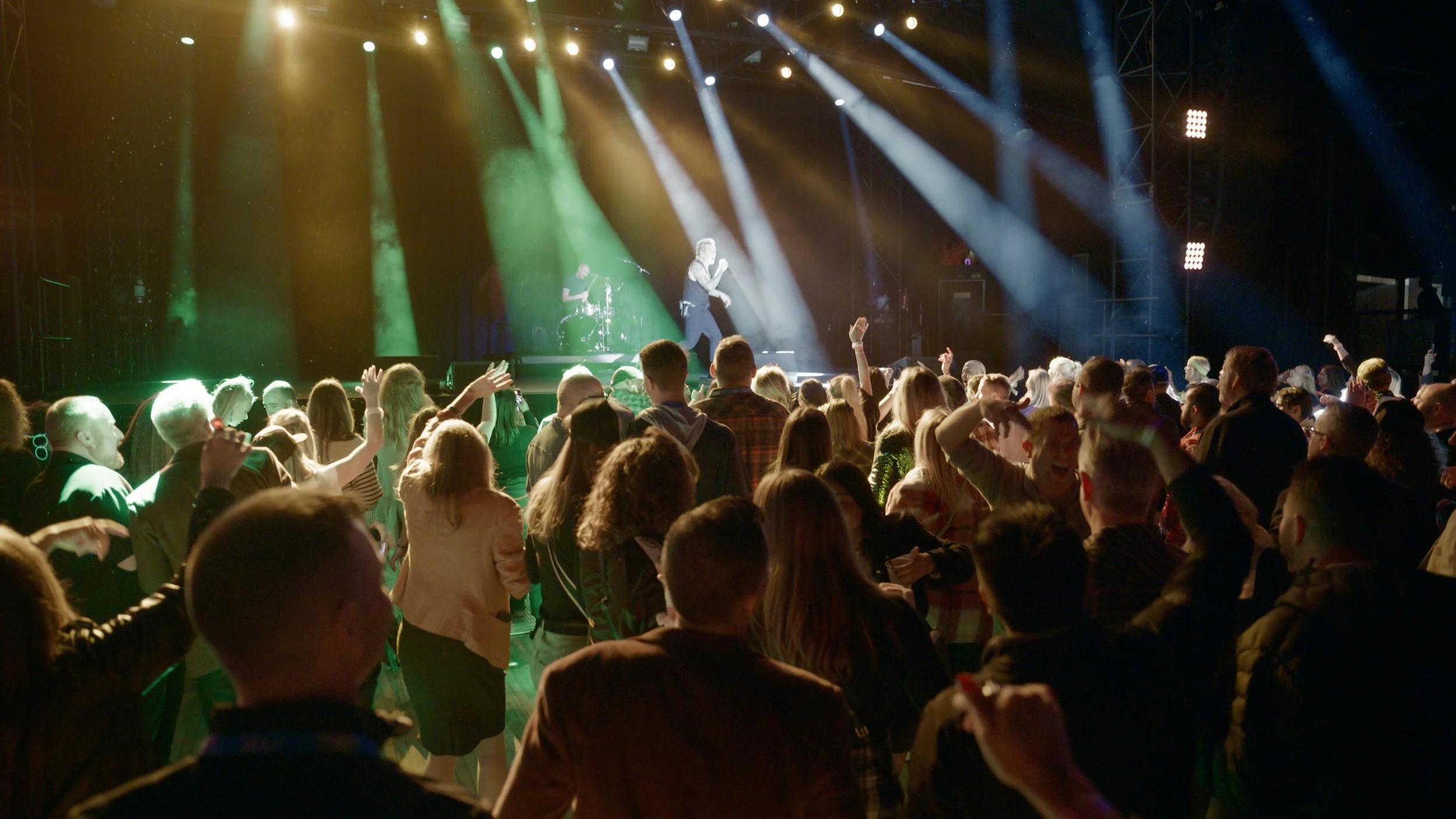 A concert with a large crowd of people dancing and enjoying a live performance on stage. The stage is brightly lit with colorful spotlights, and a singer is performing with a band in the background.