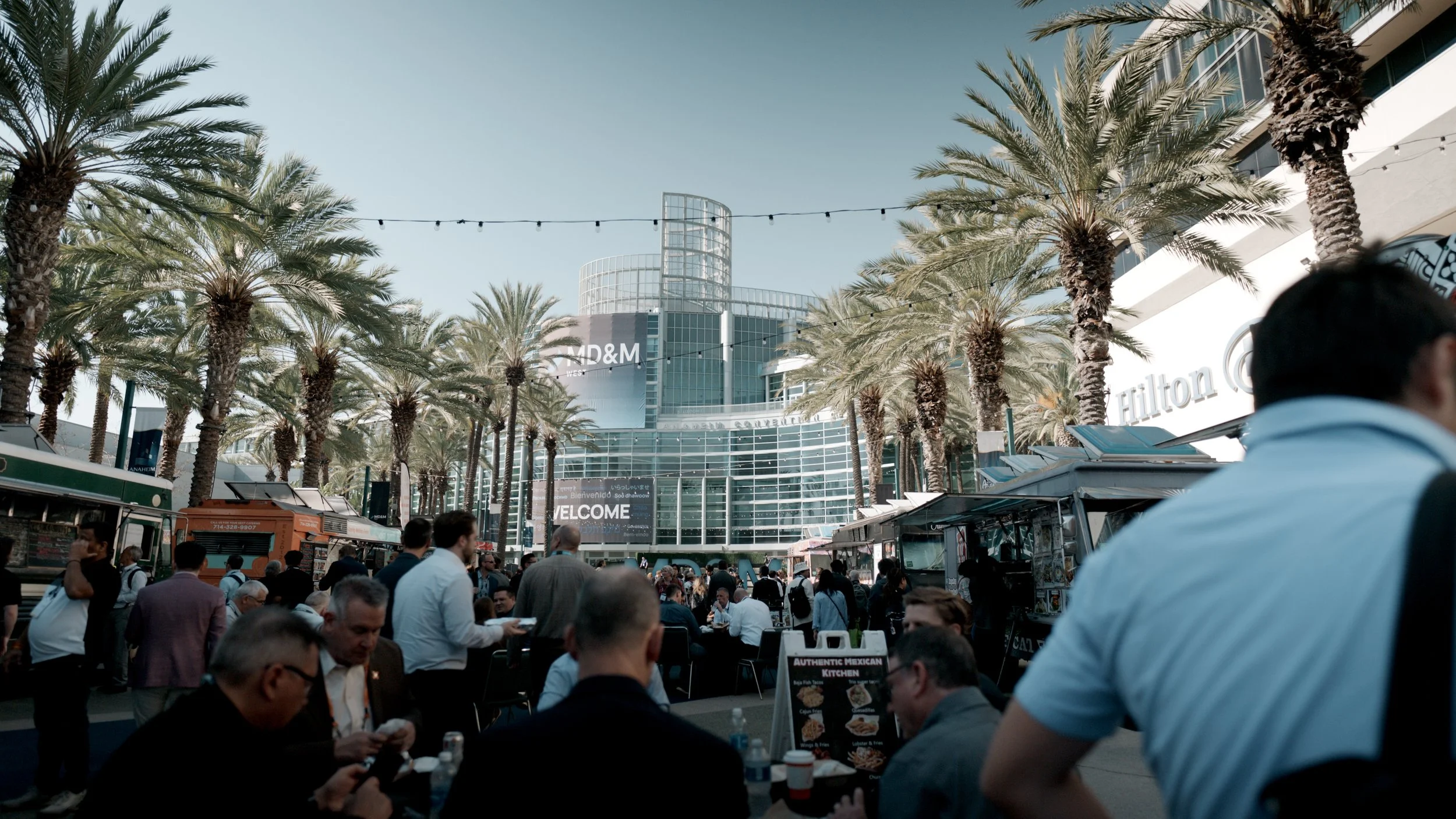 Crowd of people gathered outdoors near a modern glass building with palm trees and string lights overhead.