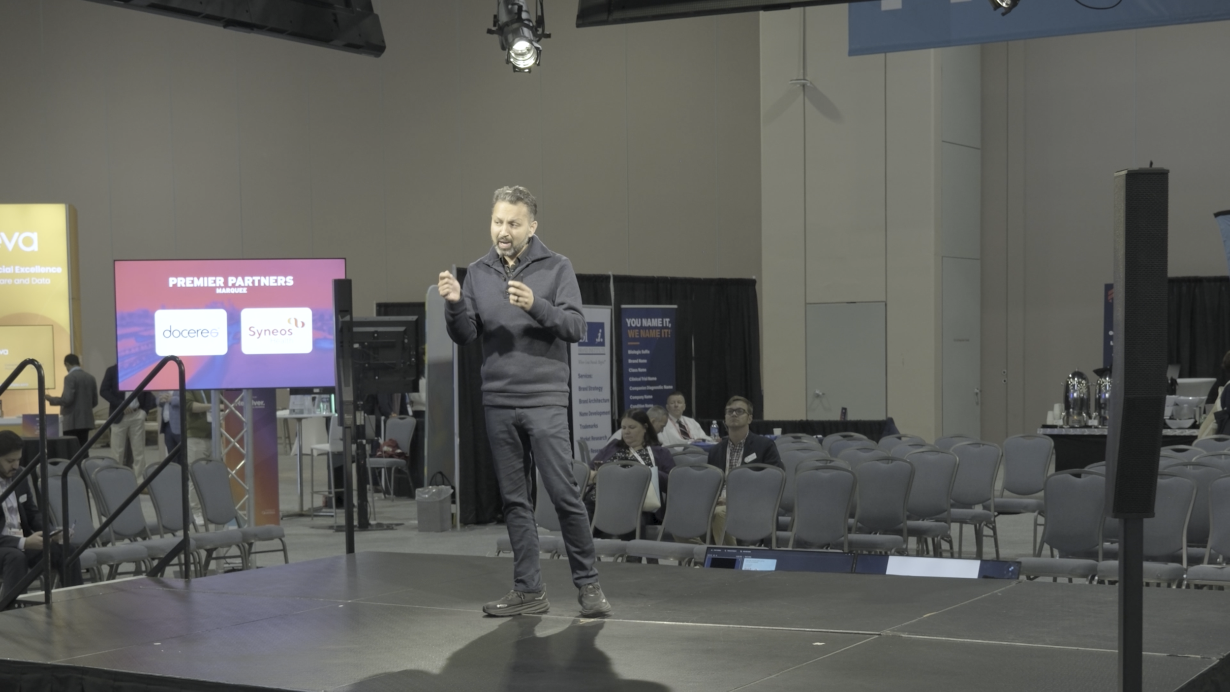 A man in a gray jacket and dark pants giving a presentation on a stage at a conference. There are multiple screens, chairs, and attendees in the background.
