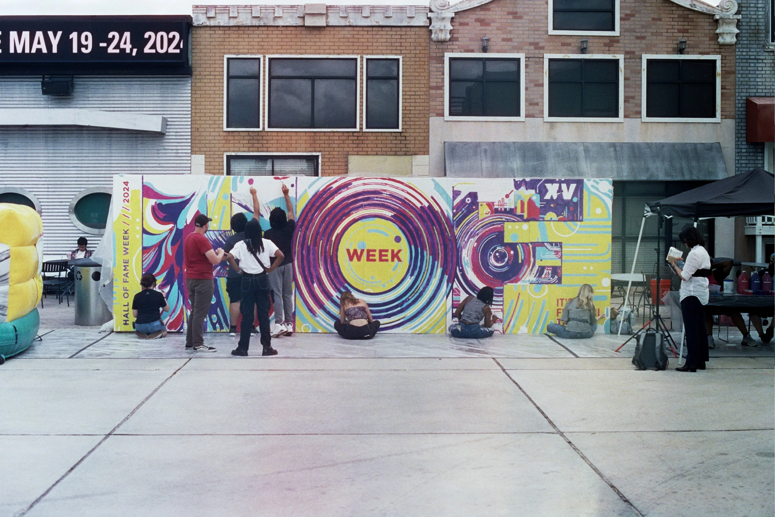 People gathered in front of a colorful mural at an outdoor event, with some sitting and others standing, and a woman reading a book to the group. There are tents and brick buildings in the background.