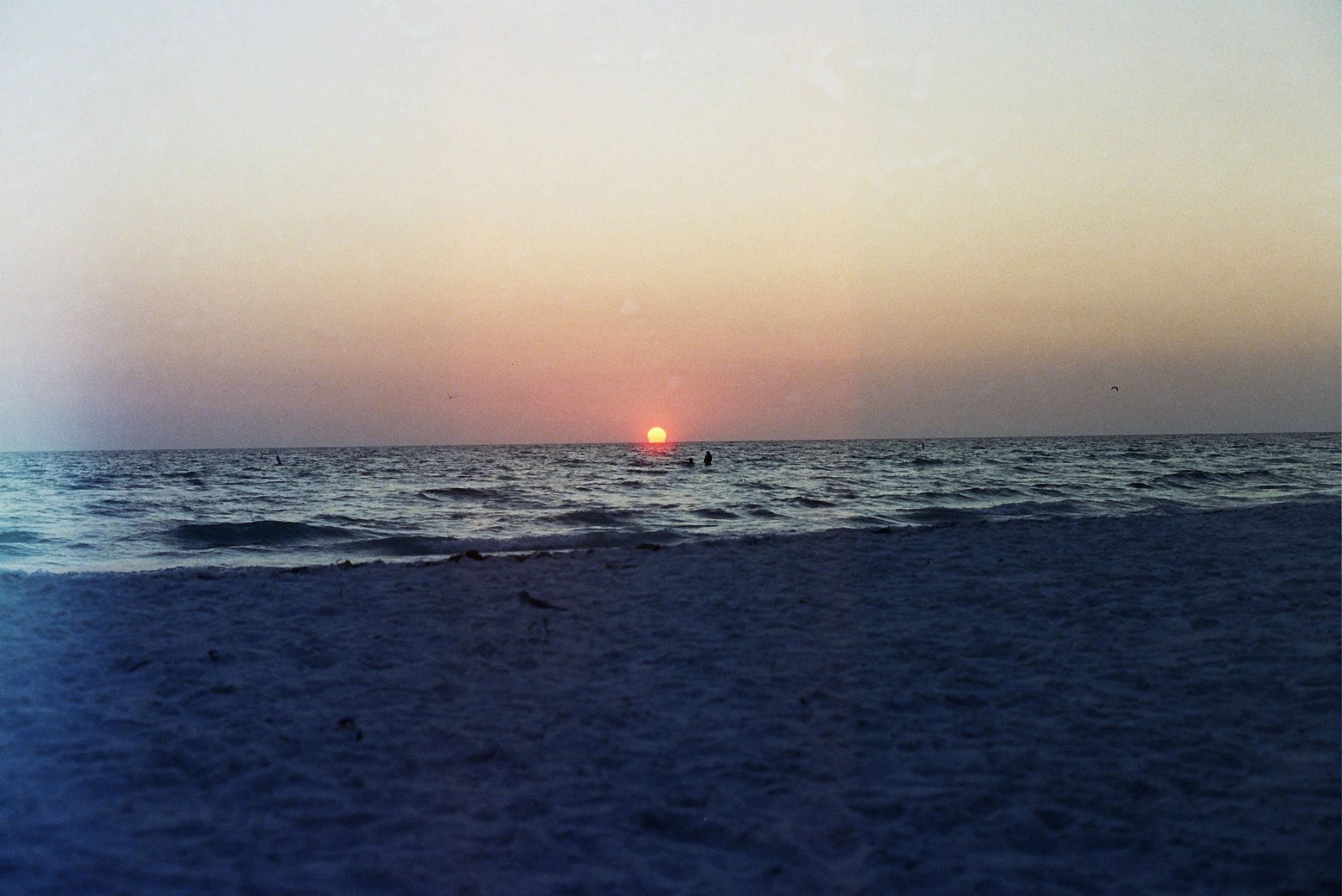 Sunset over the ocean seen from the beach with a few people in the water and a peaceful sky.