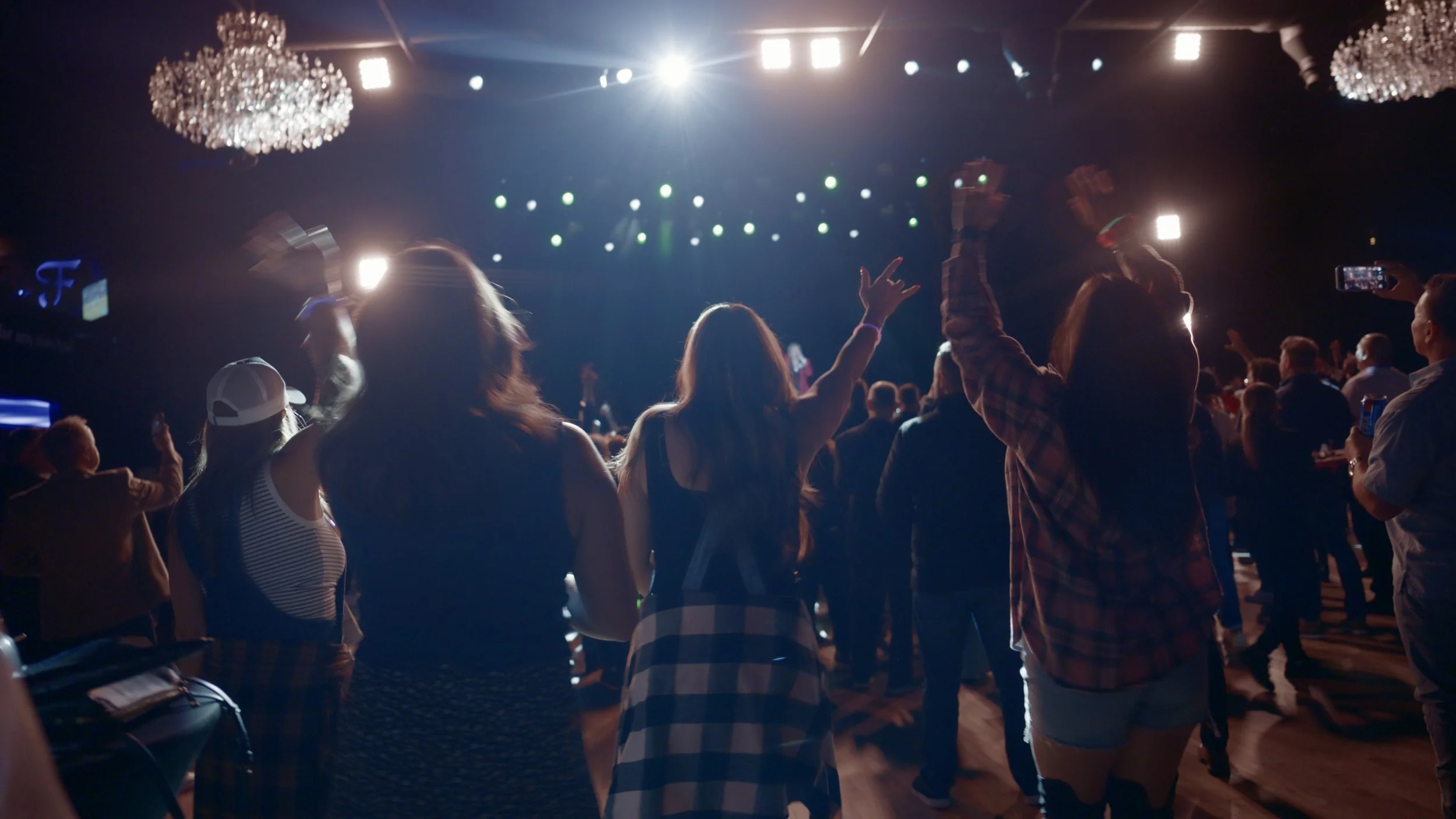Crowd of people dancing and enjoying a live concert or performance on a stage with bright lights and chandeliers overhead.