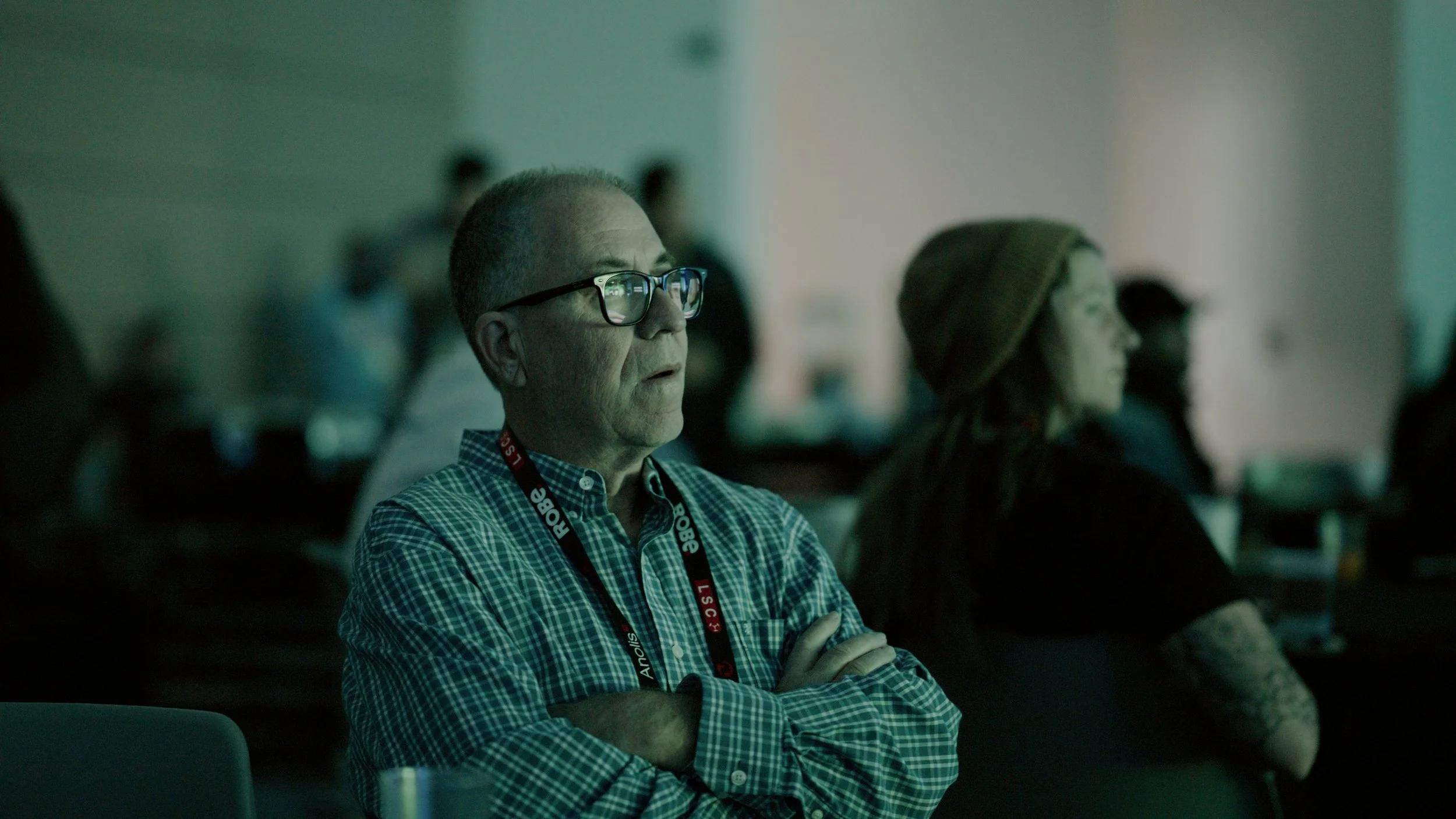 A man with glasses and a checked shirt attending a conference or presentation, with other attendees visible in the background.