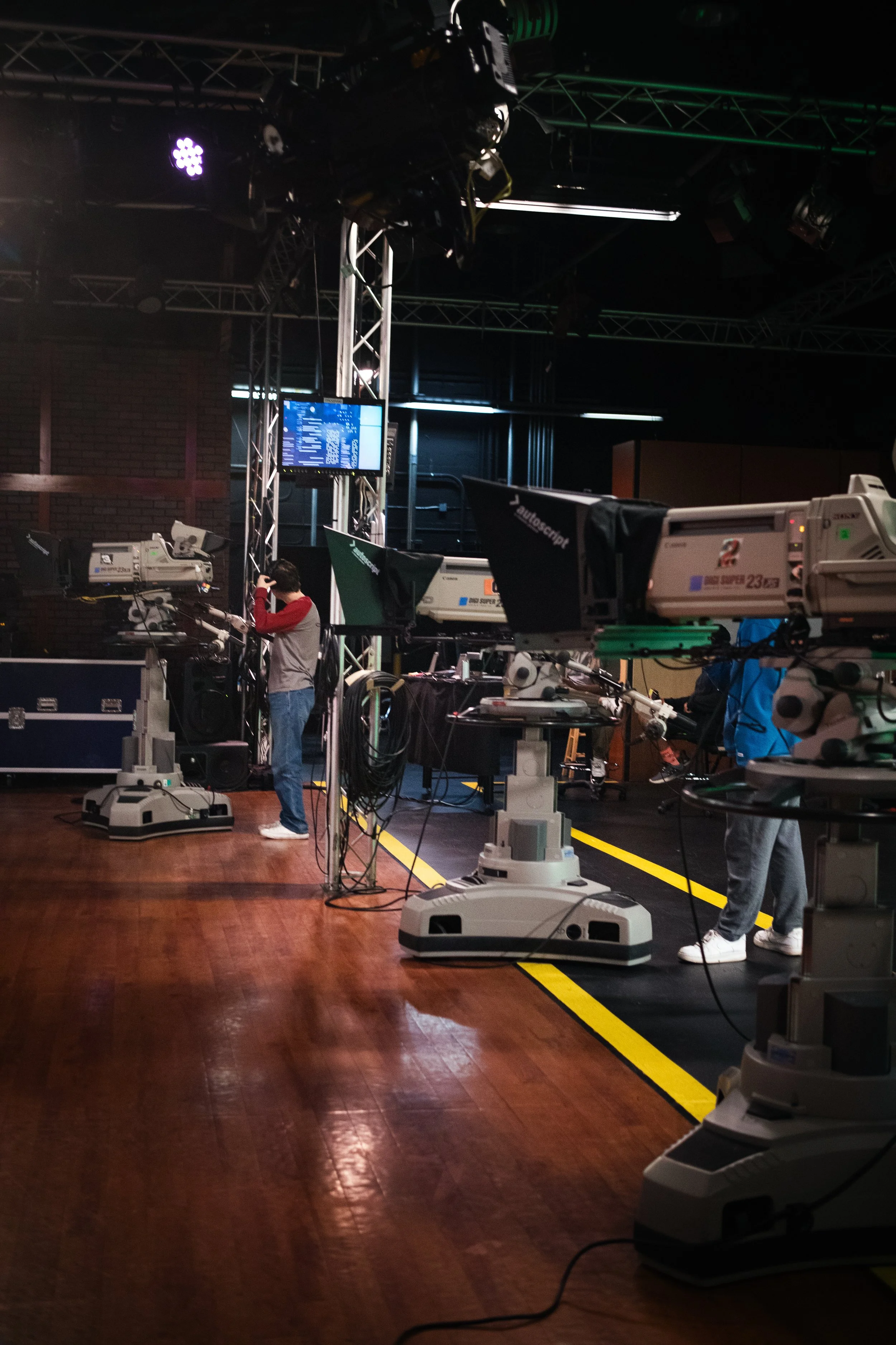 Television studio with cameras and crew setting up, dark room with industrial lighting and equipment.