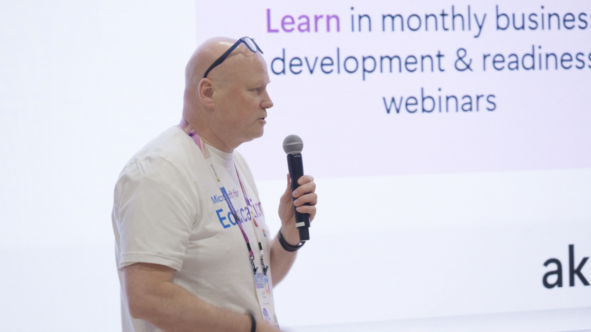 A bald man wearing glasses on his head and a white t-shirt with event-related text, holding a microphone and speaking at a conference or seminar. There is a large screen behind him displaying text about learning, monthly business development, and webinars.