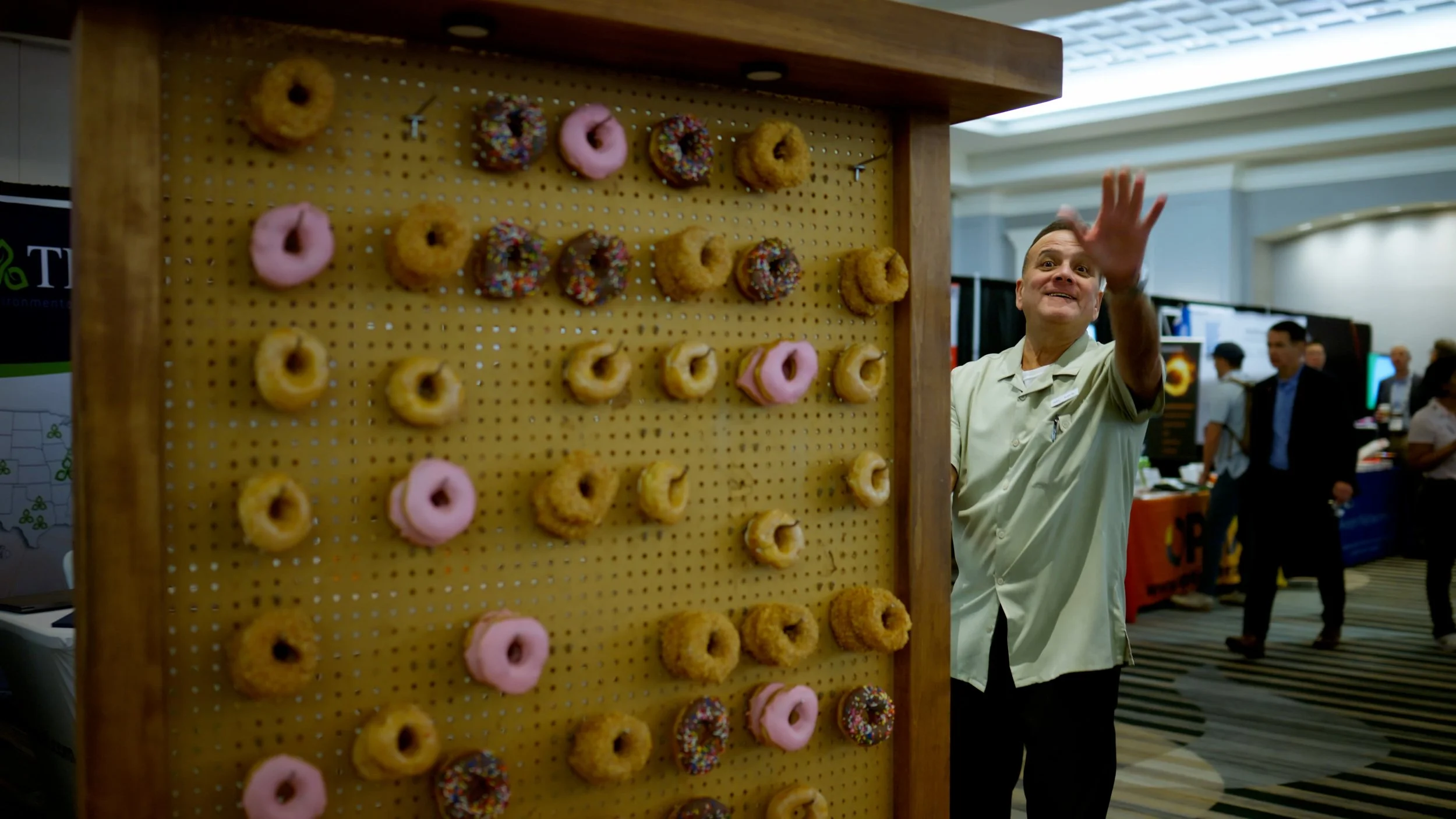 A display of assorted donuts on a pegboard, with pink frosted, chocolate frosted, and sprinkled varieties, at an indoor event. A man in a white shirt is reaching out or waving nearby, with several people in the background.