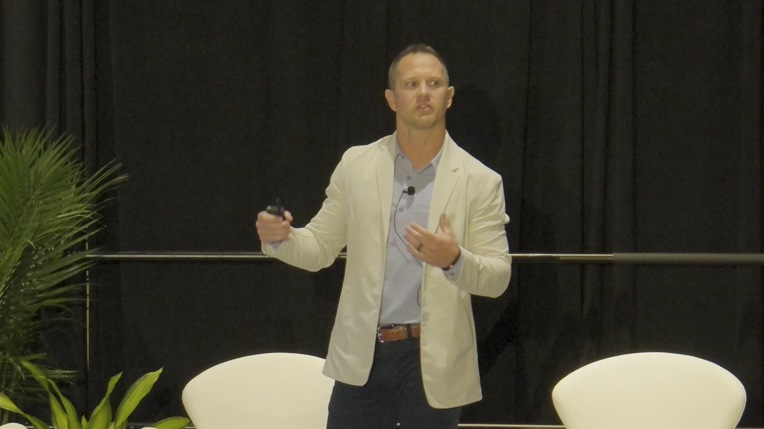 A man giving a presentation in a conference room with black curtains, white chairs, and some green plants visible on the side.