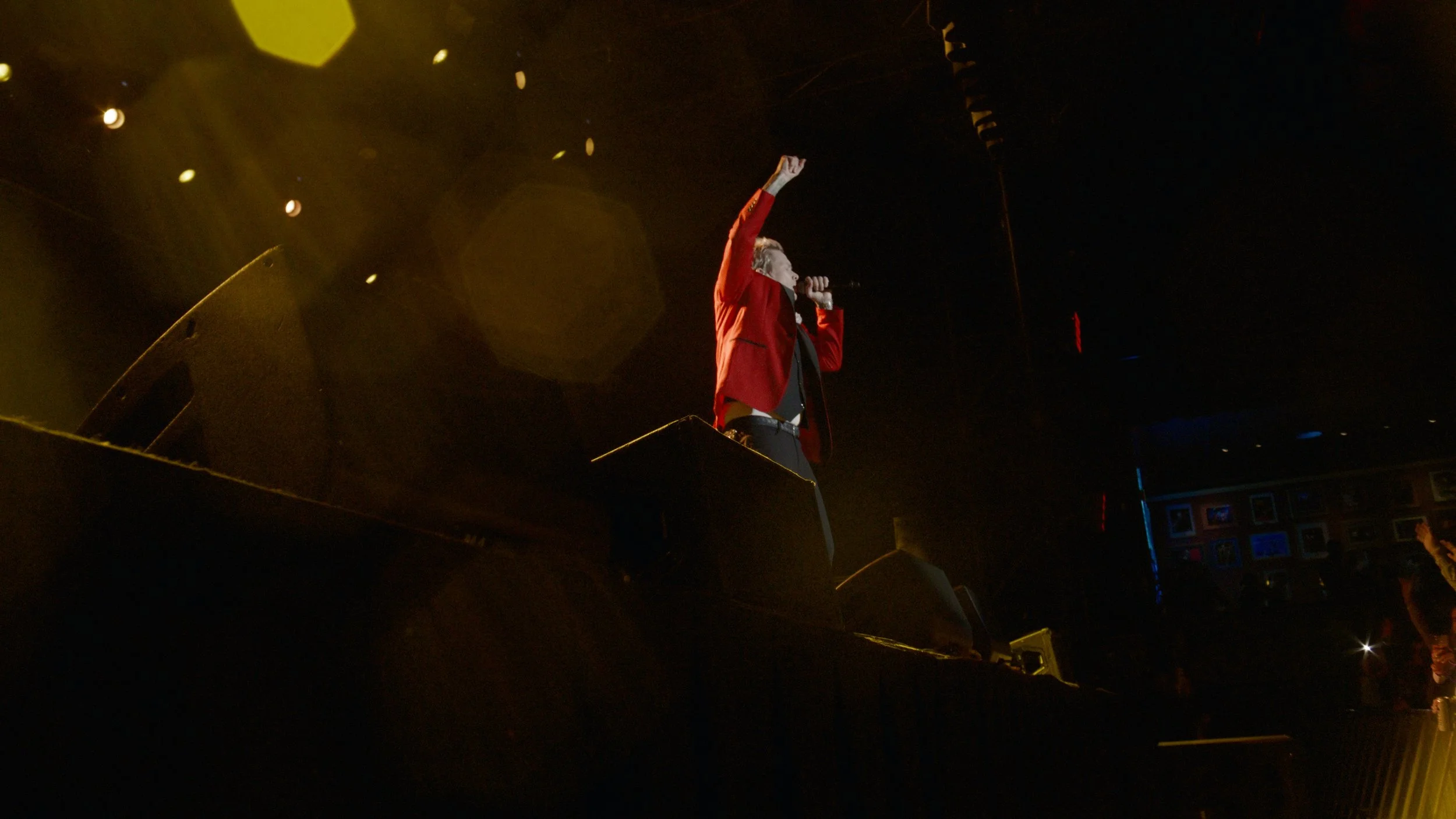 A person in a red jacket is singing into a microphone on stage, with one arm raised. The scene is dark with stage lights and a background that appears to have framed pictures.