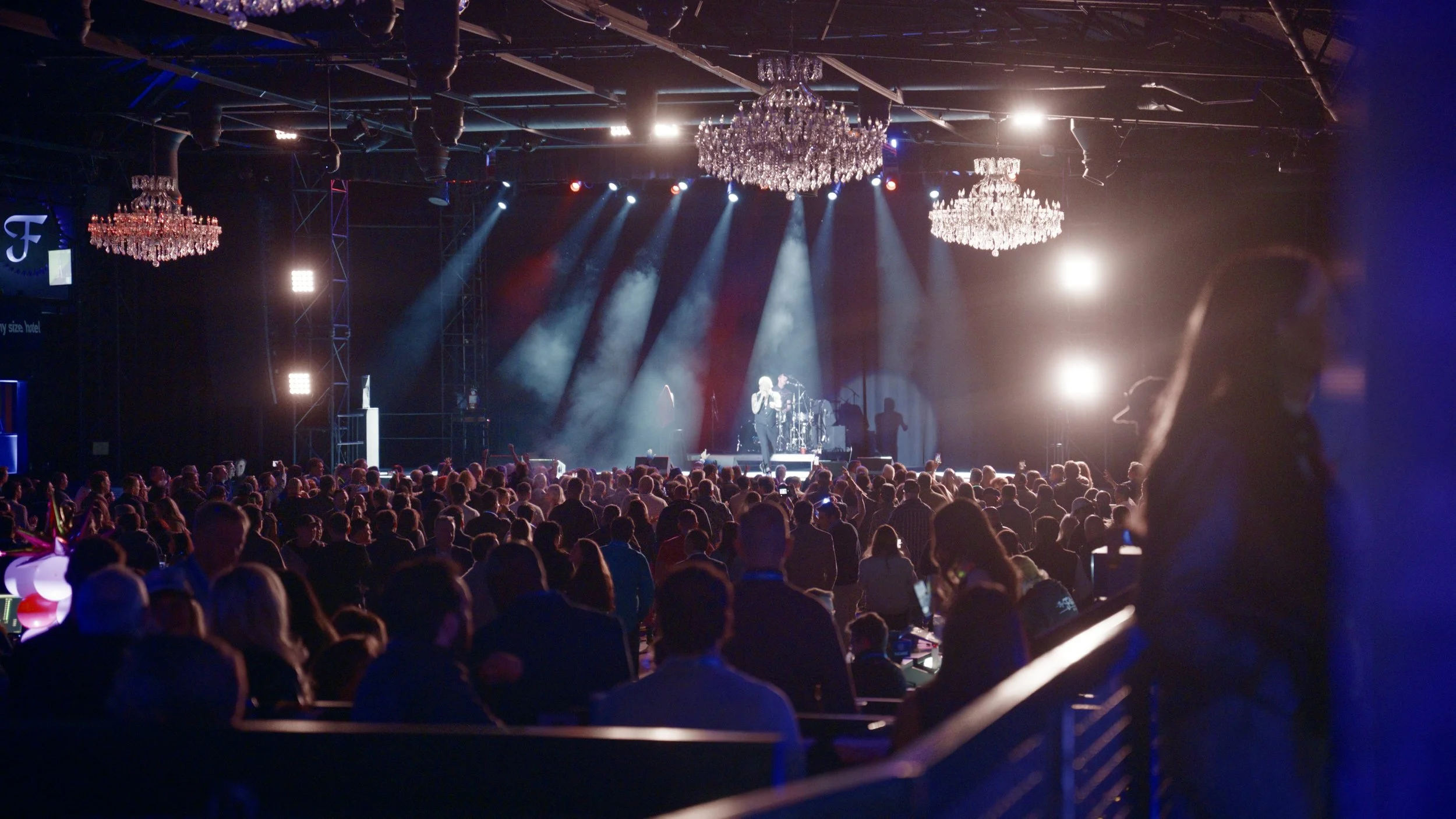 Concert hall with performer on stage and large audience, illuminated chandeliers hanging from ceiling, stage lights shining on performer.