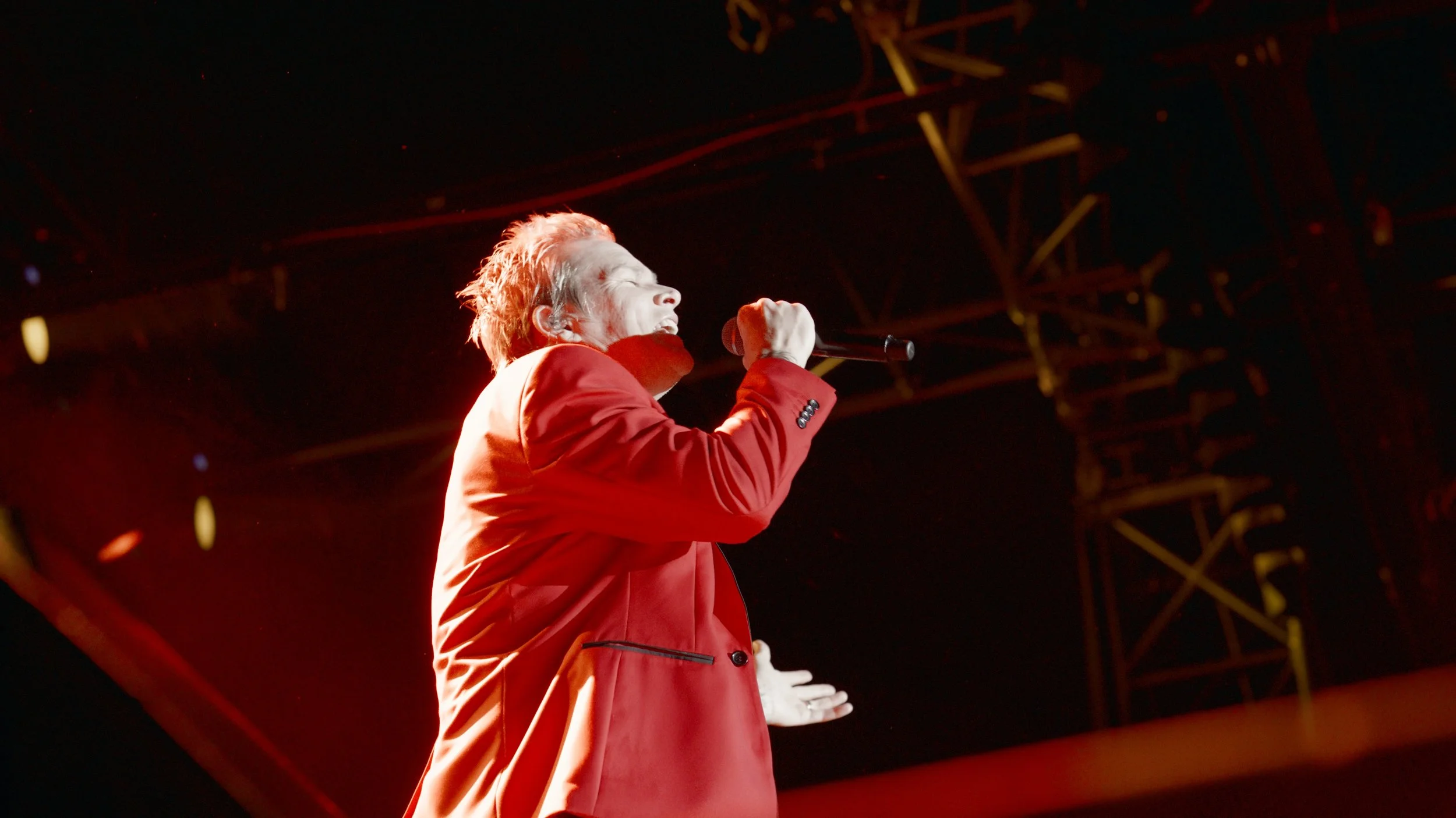 A person singing into a microphone on stage, wearing a red jacket, with a dark stage background and stage lighting.