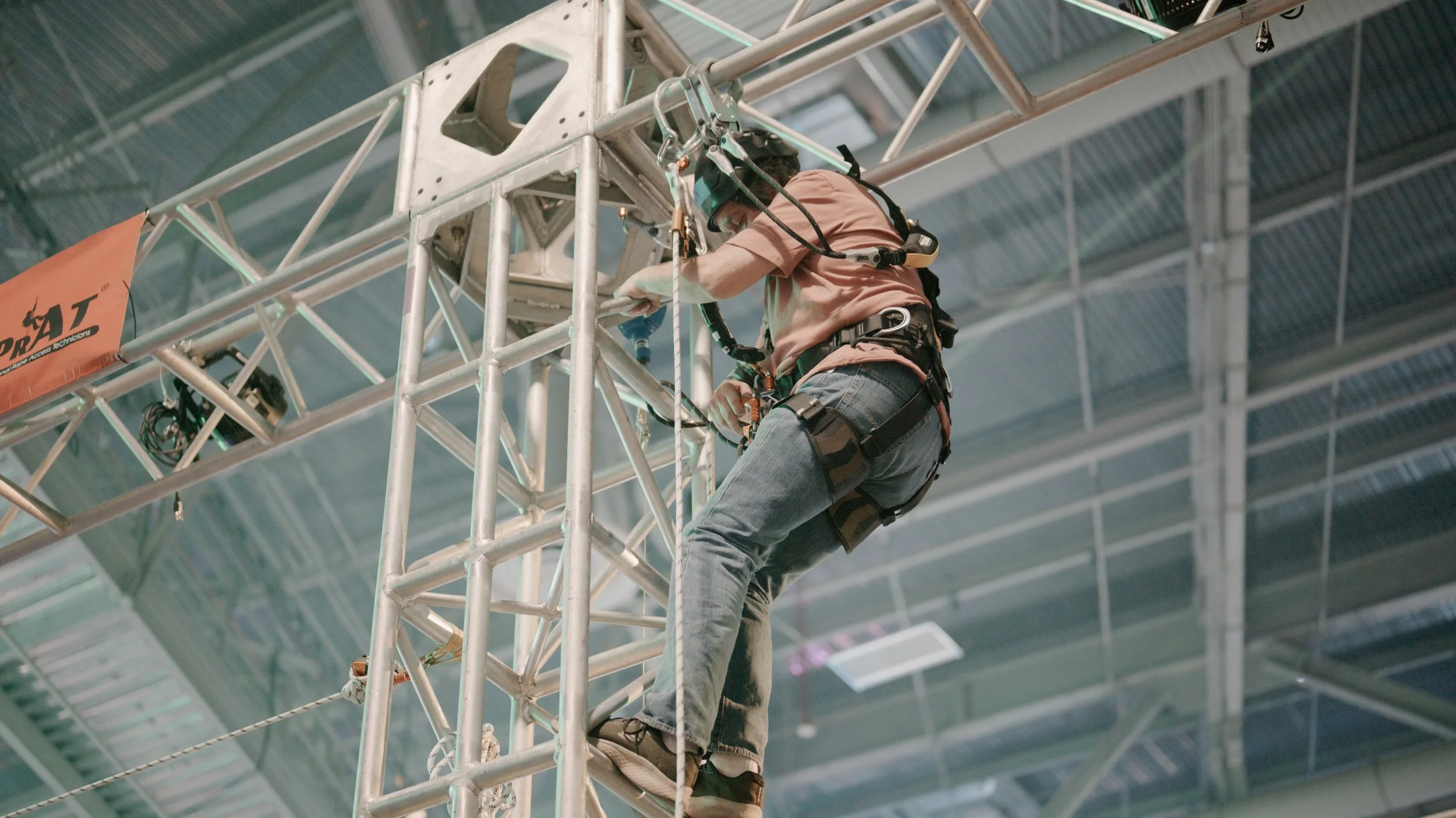 A person wearing a safety harness working on a metal truss structure indoors.