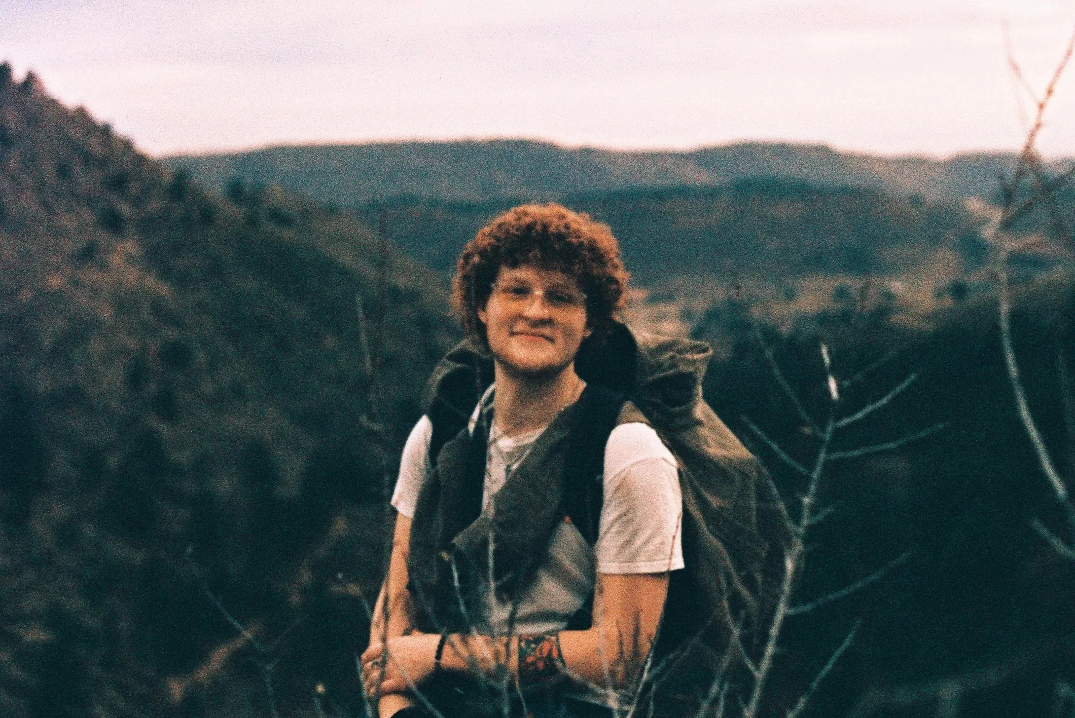 Person with curly hair smiling while riding a bike through a wooded area during dusk or dawn.