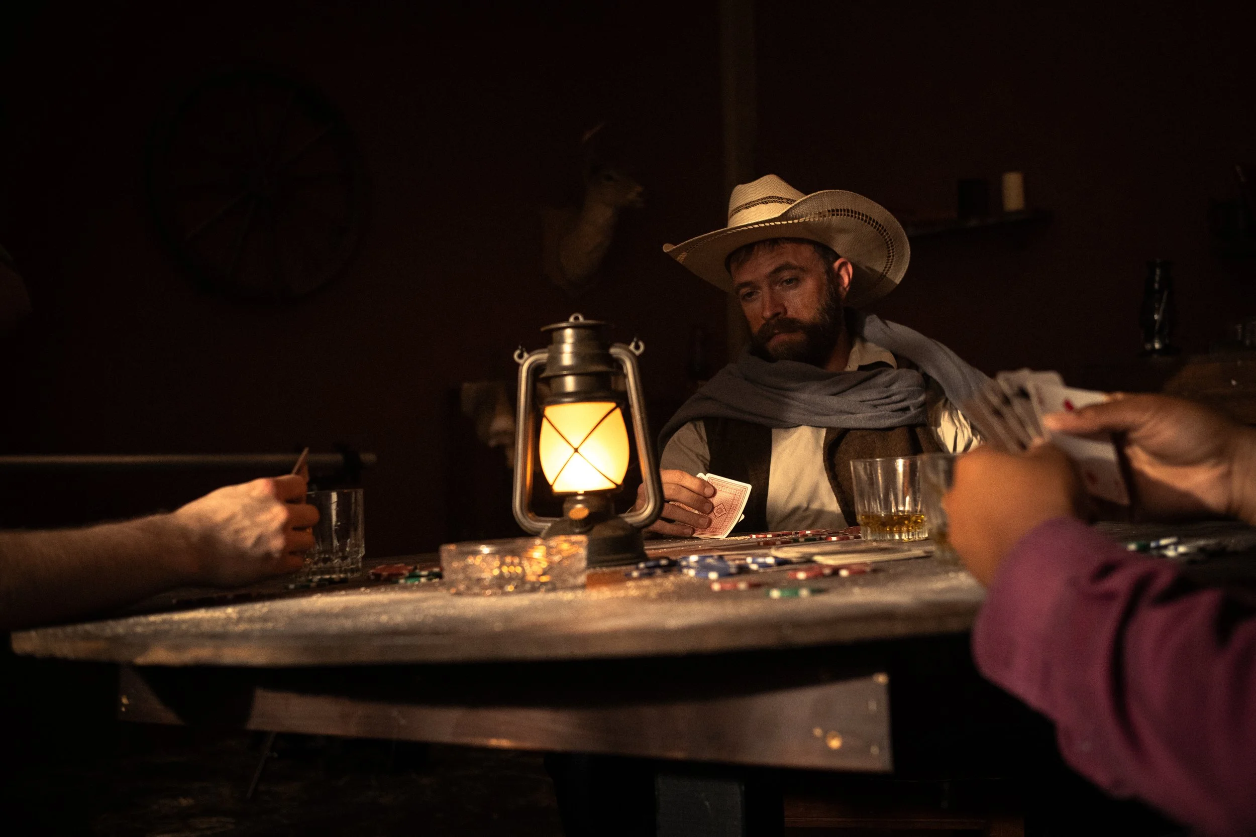A man wearing a cowboy hat playing cards at a dimly lit table with poker chips and glasses, with a lantern providing light, in a western-themed setting.