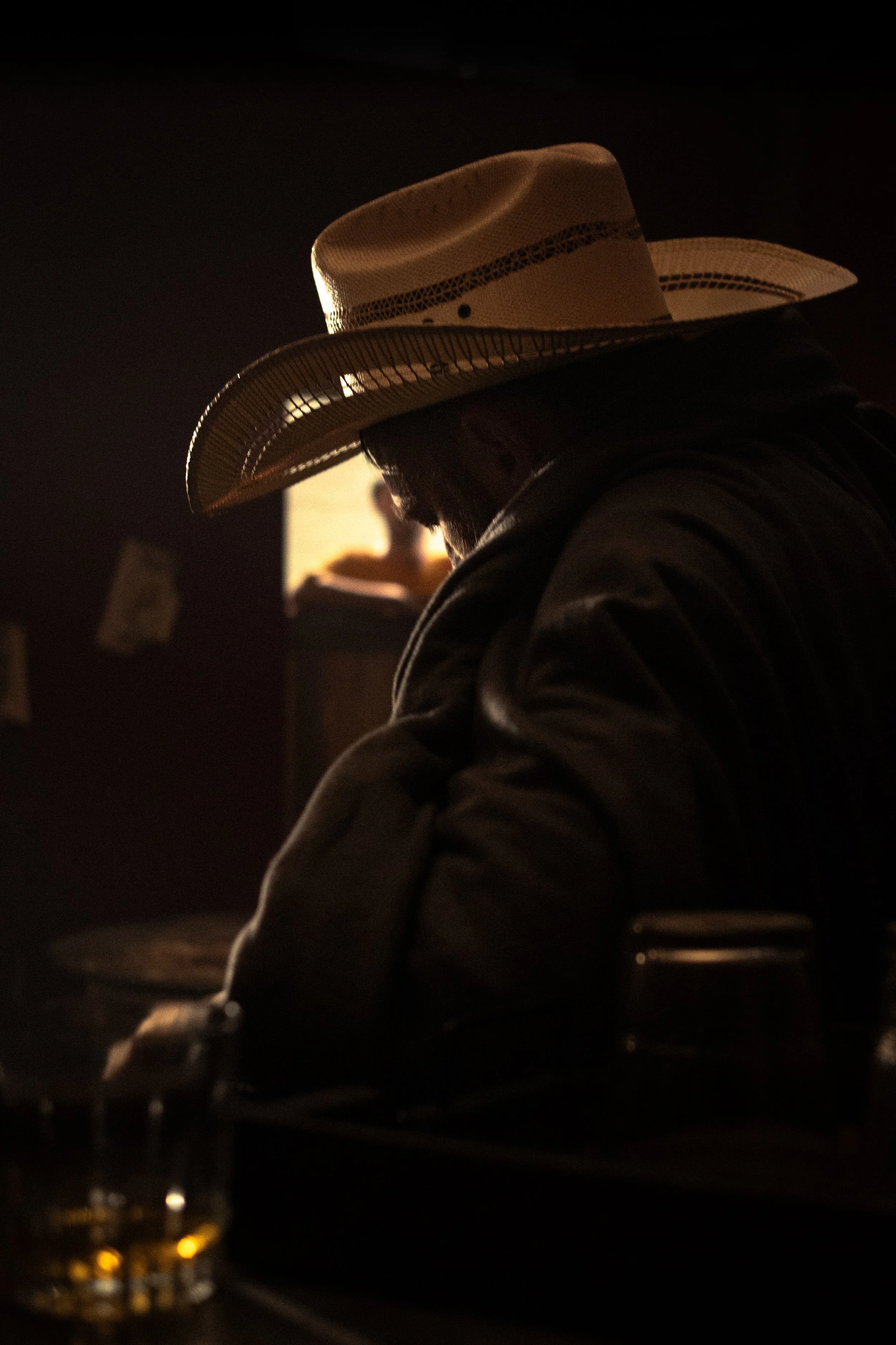 Man in a cowboy hat sitting at a table with a drink in a dimly lit setting, possibly a bar or restaurant, with a blurred figure in the background.