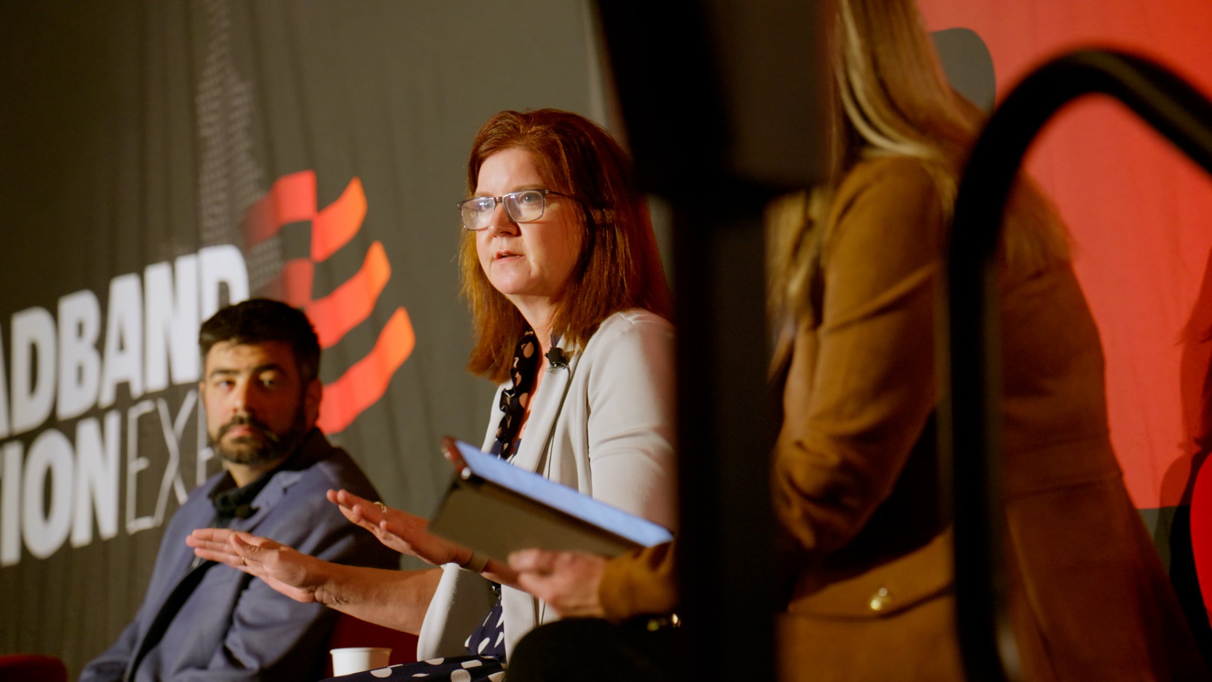 A woman with red hair, glasses, and a white blazer speaking at a panel discussion, with a man with dark hair and a beard seated next to her, and other panelists partially visible.