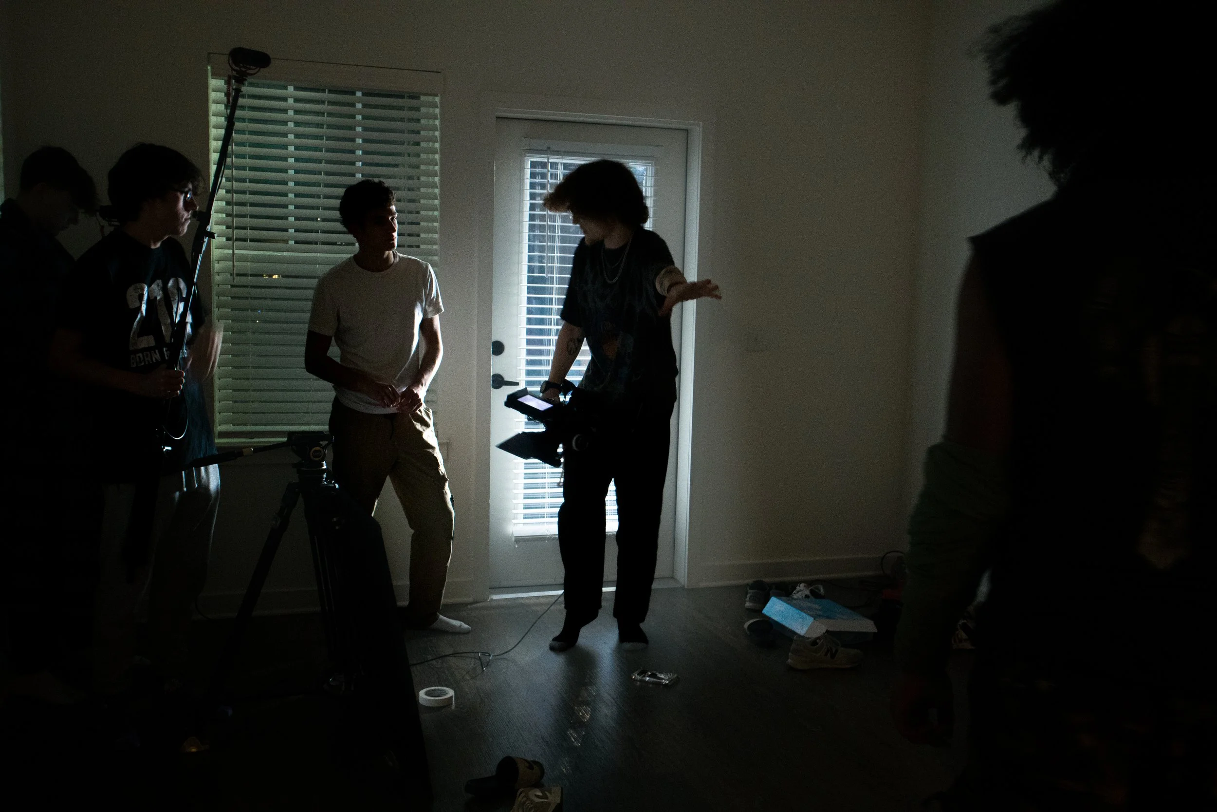 A group of young people are in a dimly lit room, some engaging in conversation and others standing around, with a door with blinds and a window in the background.