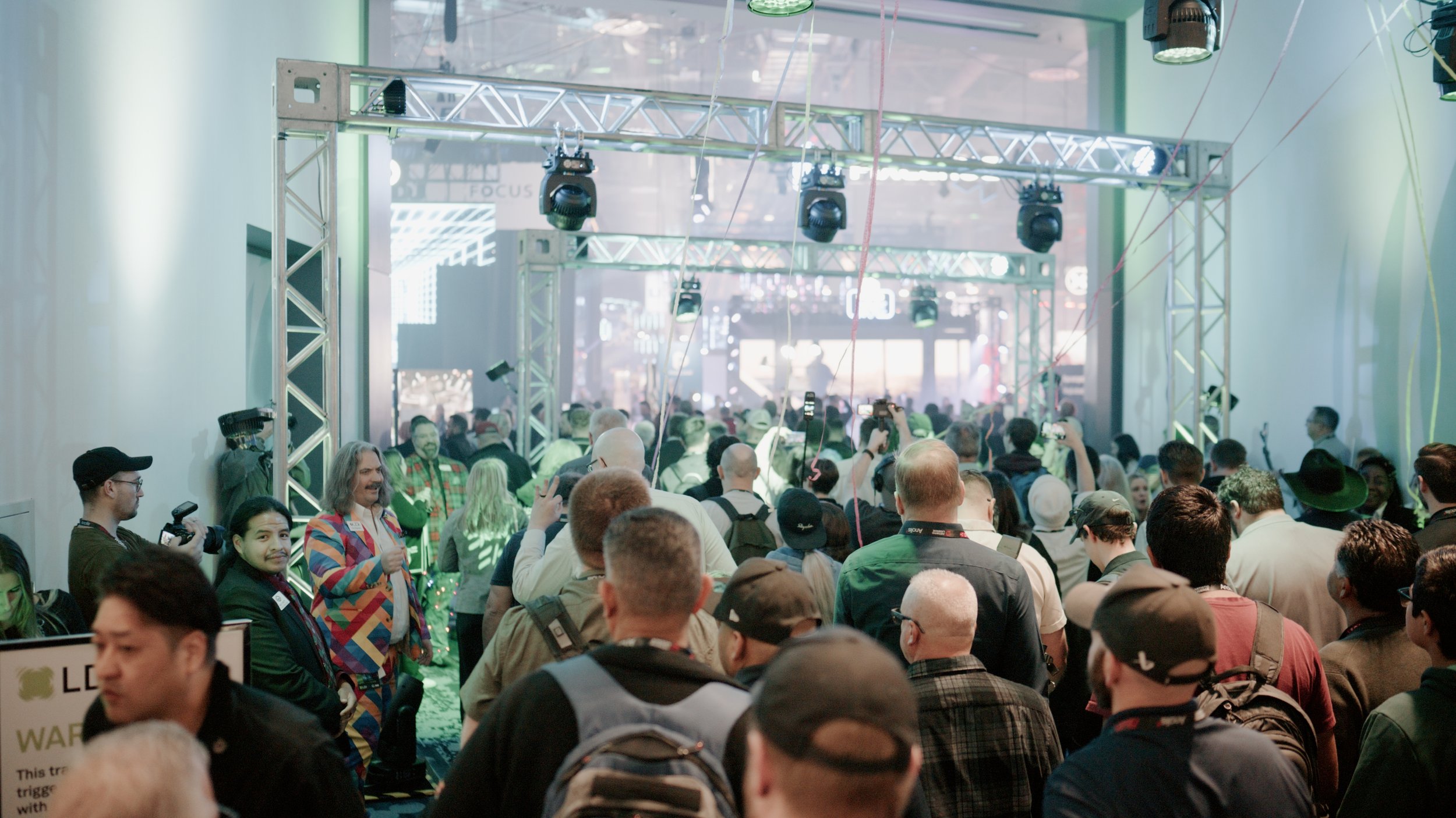 A crowded indoor event with many people gathered, some taking photos or videos, under a structure with stage lights, with a large screen or display in the background.