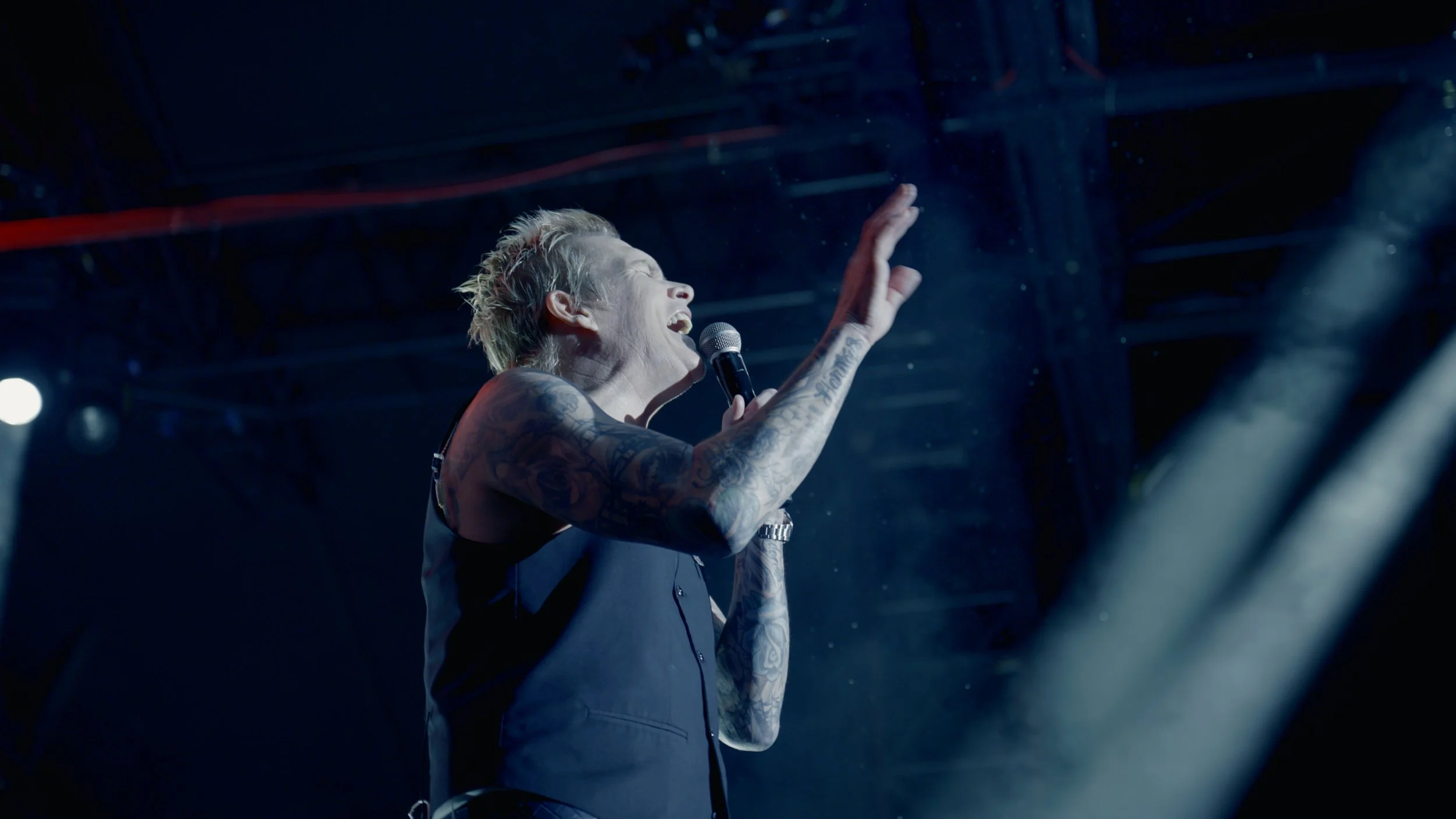 A woman with short blonde hair singing into a microphone on stage, wearing a dark vest and showing tattoos on her arms, with stage lights in the background.