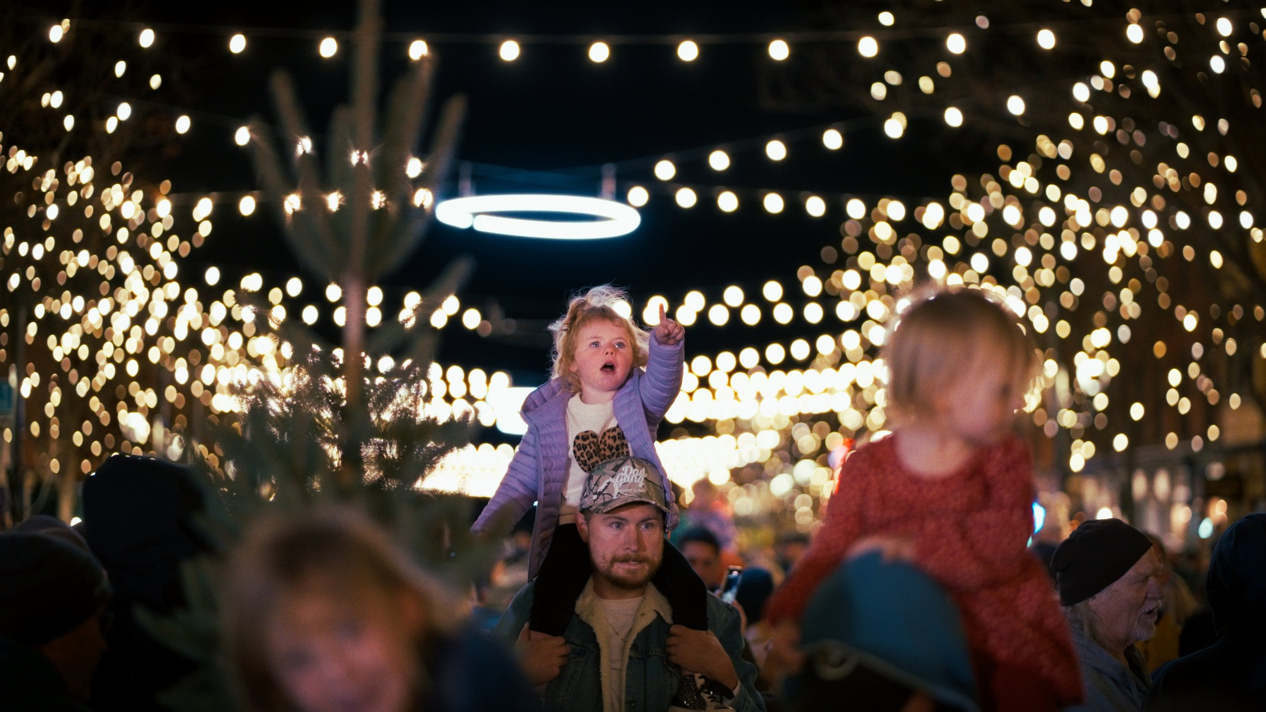 Children and adults gathered outdoors at night, illuminated by string lights, with a young girl pointing upward on an adult's shoulders, festive atmosphere.