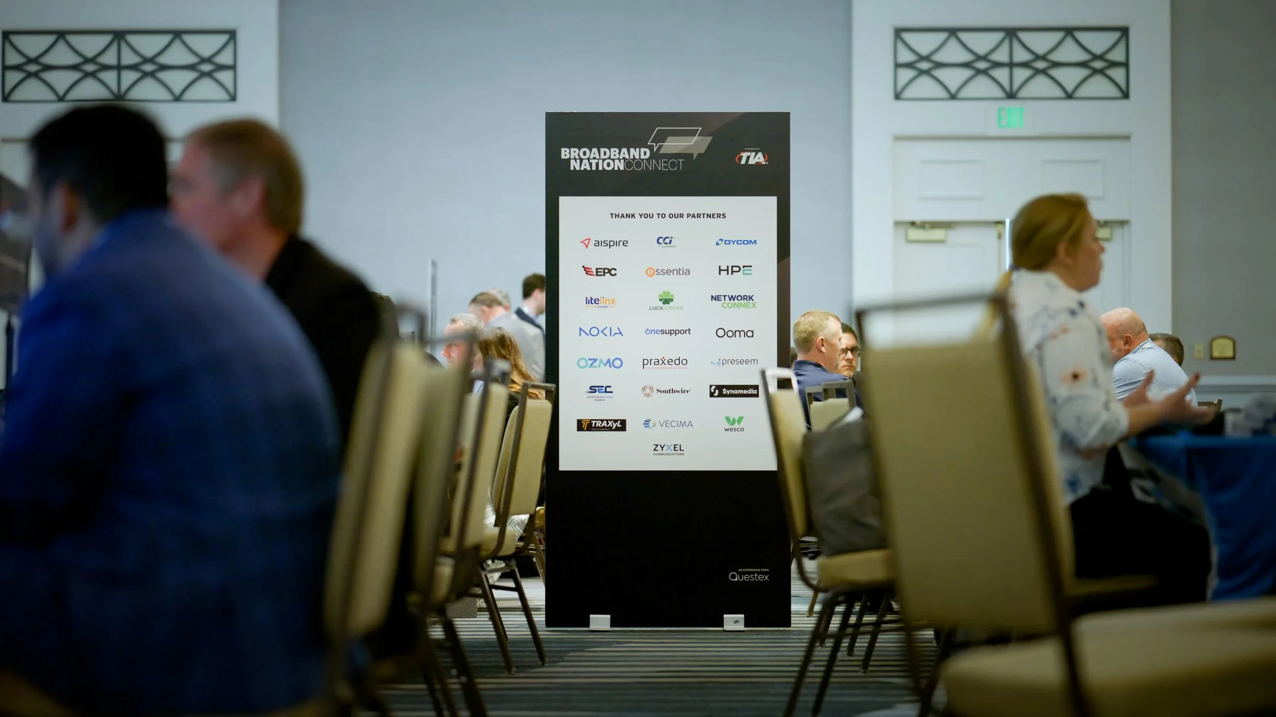 A conference room with a large vertical sign featuring logos of various companies and the text 'Broadband Nation Connect'. Attendees are seated in rows, having conversations and working, with some blurred figures in the foreground and background. The