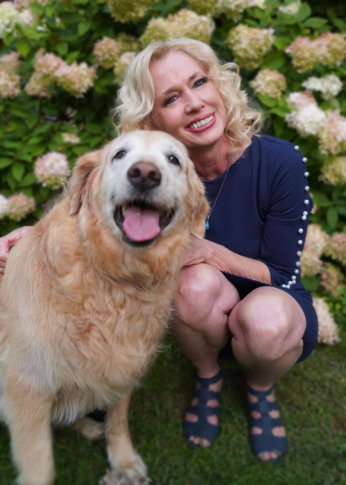 A woman with blonde curly hair smiling next to a happy golden retriever in front of a bush with pink and white hydrangea flowers.
