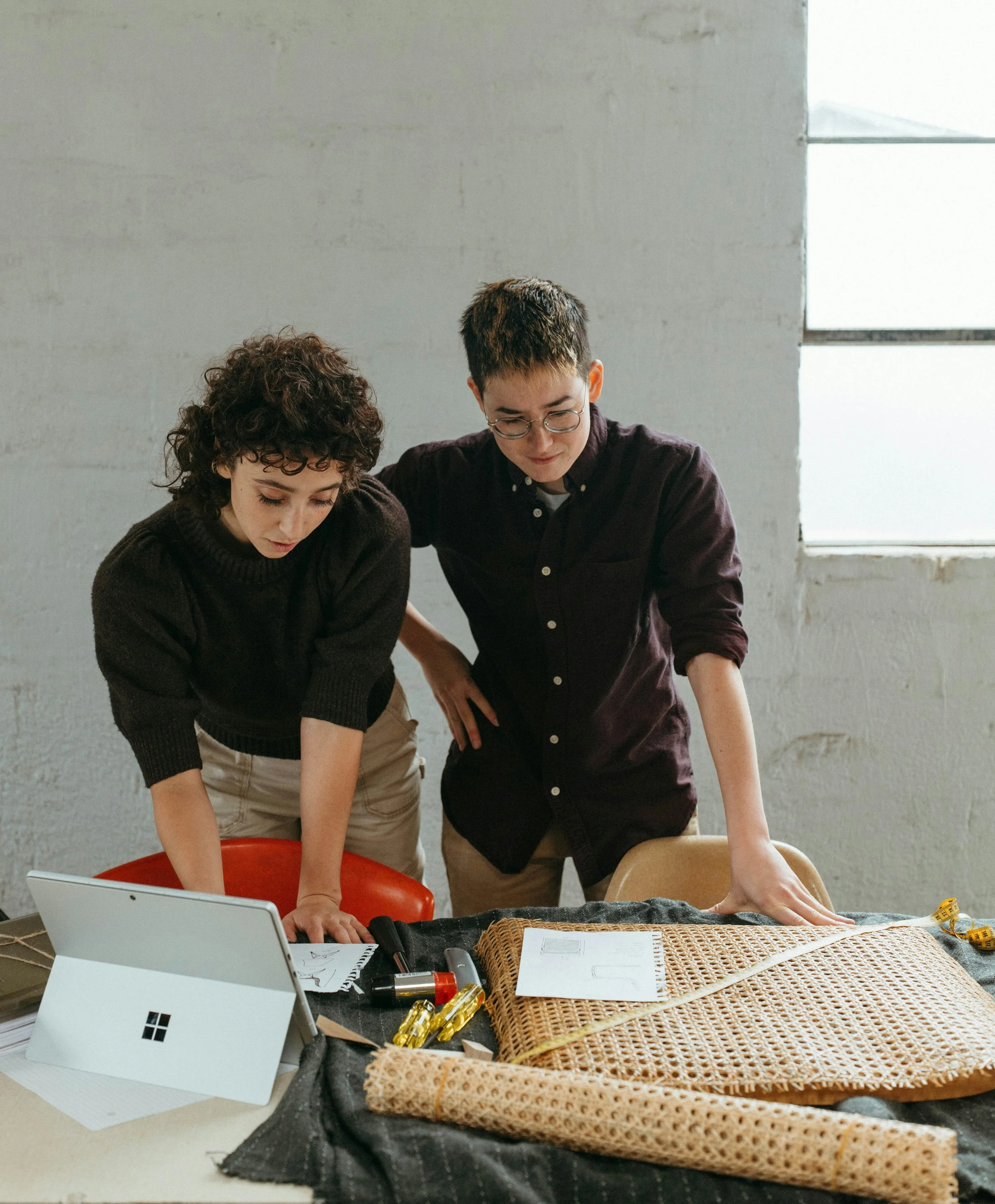 Two queer individuals leaning over a table working together on a creative project.