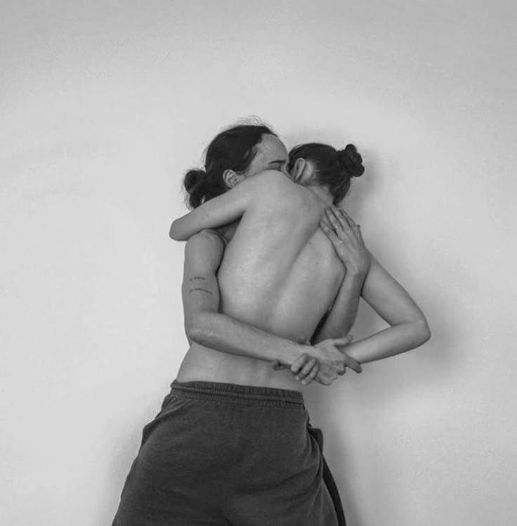 Black and white photo of an adult and a child hugging each other, both topless, against a plain wall.