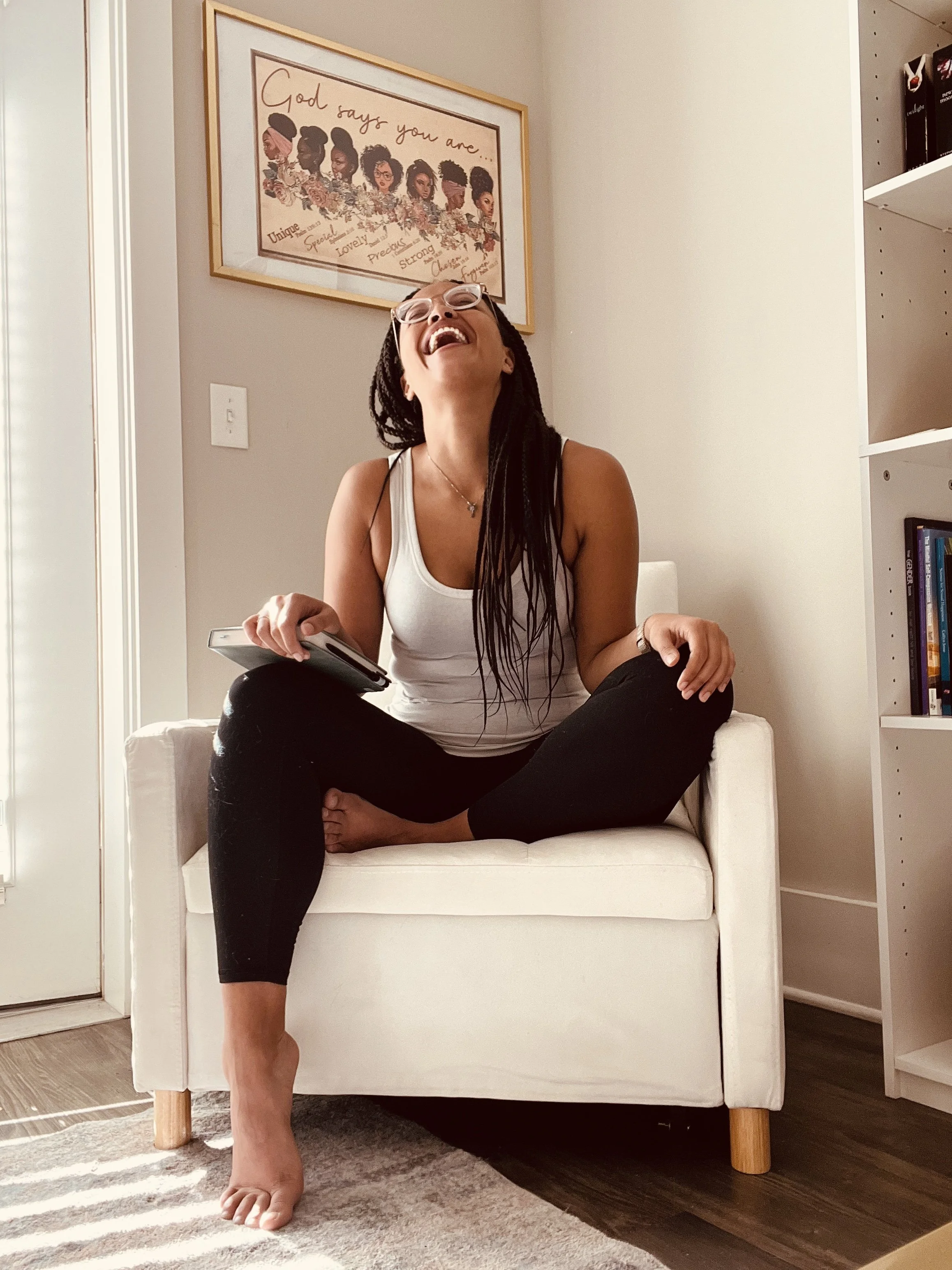 A woman with glasses and dreadlocks, sitting cross-legged on a white armchair, laughing with her head tilted back, holding a remote in one hand. She is wearing a white tank top and black pants. There is a framed picture on the wall behind her with illustrations of women and the words 'God says you are...' above.