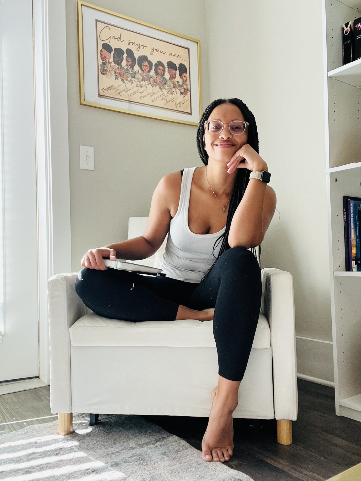A woman with glasses and long dark braided hair sitting cross-legged on a white armchair, smiling and holding a closed laptop, in a bright room with a framed artwork on the wall behind her.