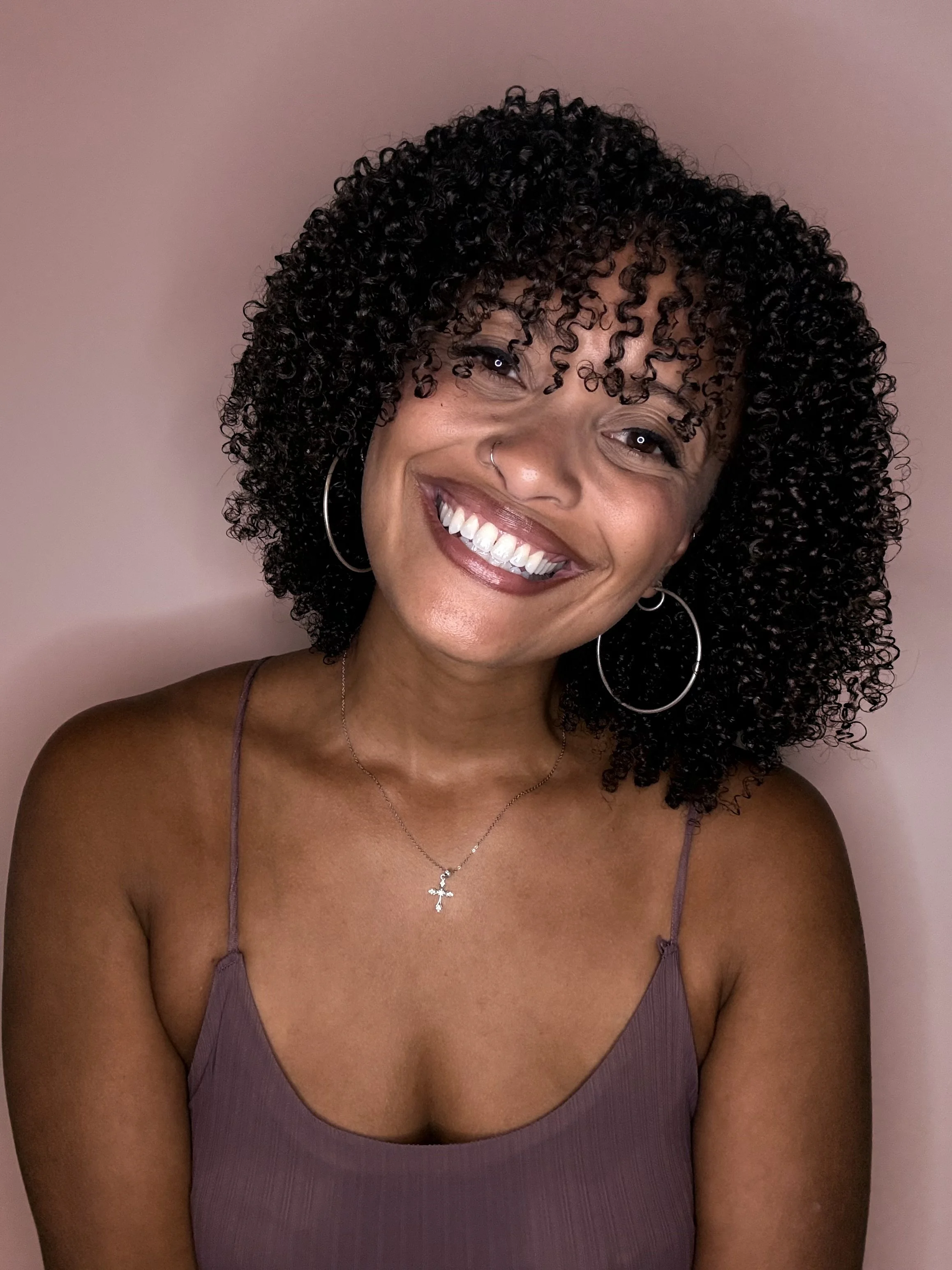 Smiling woman with curly hair, wearing hoop earrings, a necklace with a cross pendant, and a sleeveless purple top, against a pink background.
