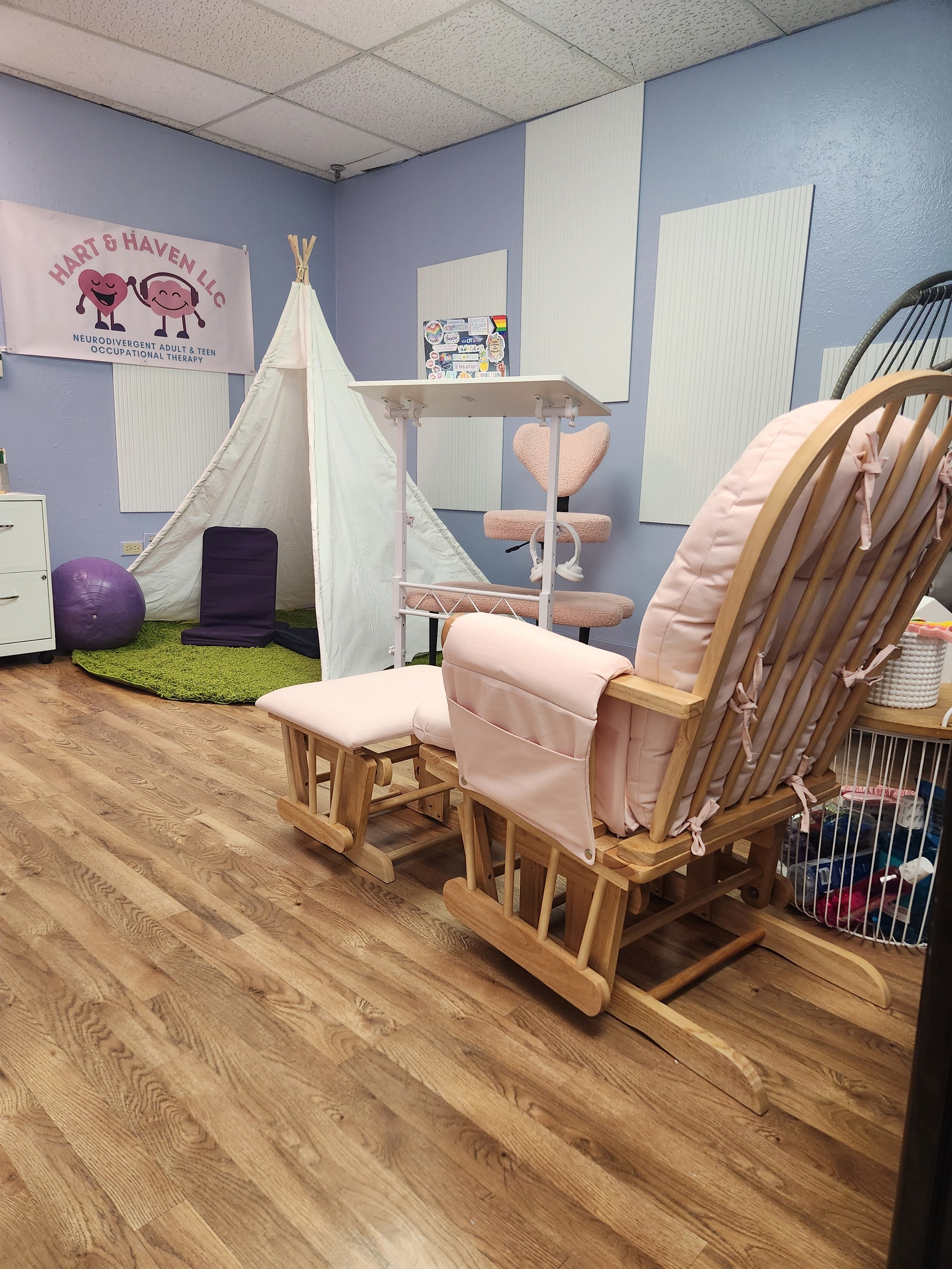 A therapy or counseling room with a wooden rocking chair, a pink padded chair with a footrest, a small table, a tent, exercise ball, and a sign for Hart & Haven LLC. The room has blue walls, acoustic panels, and a wood floor.