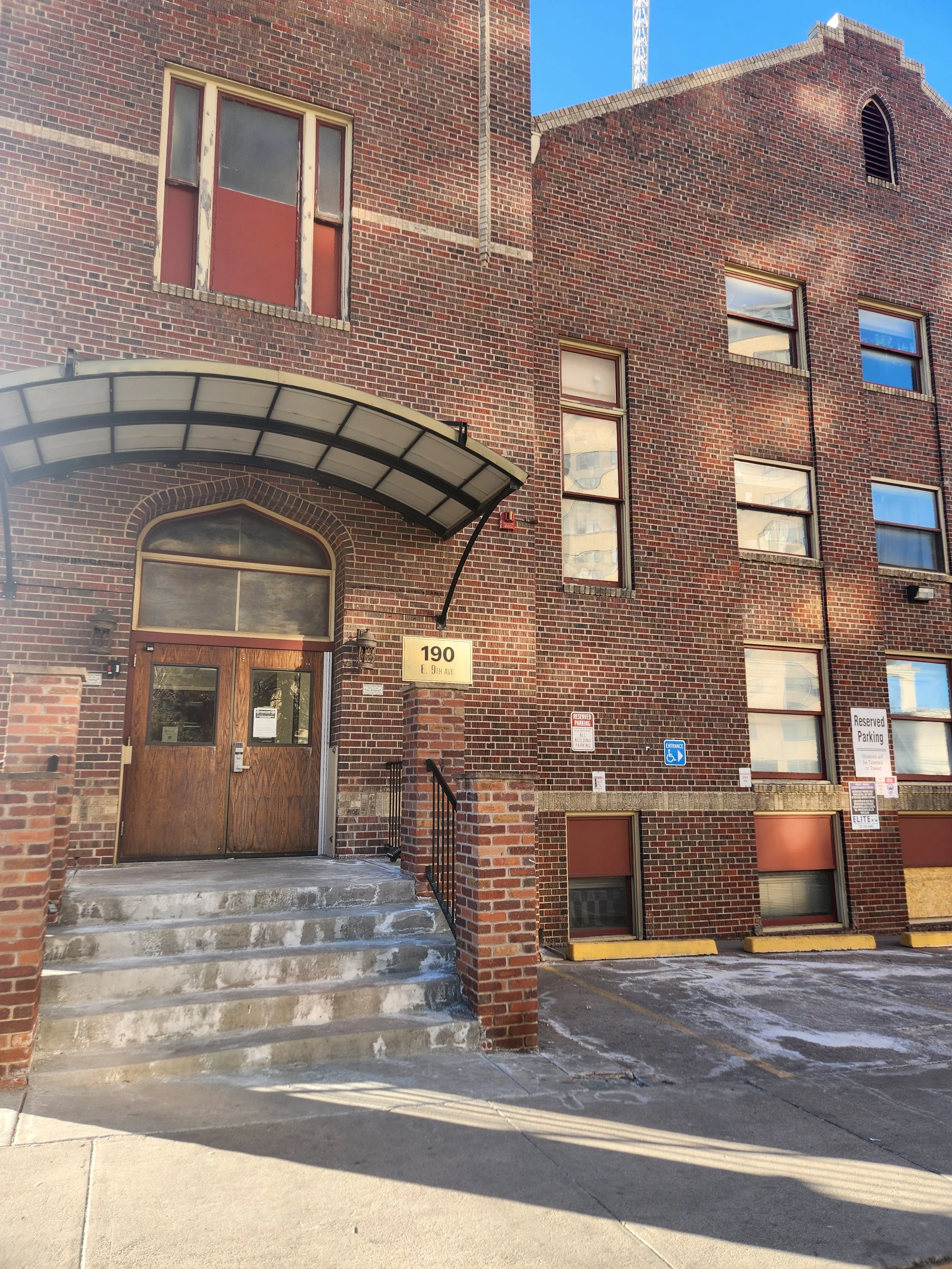 Exterior of Hart and Haven LLC office building with an arched entrance, steps, and multiple windows, some covered with red blinds, parking lot in front with yellow barriers, and blue sky overhead located at 190 East 9th Avenue, Denver CO, Unit 135
