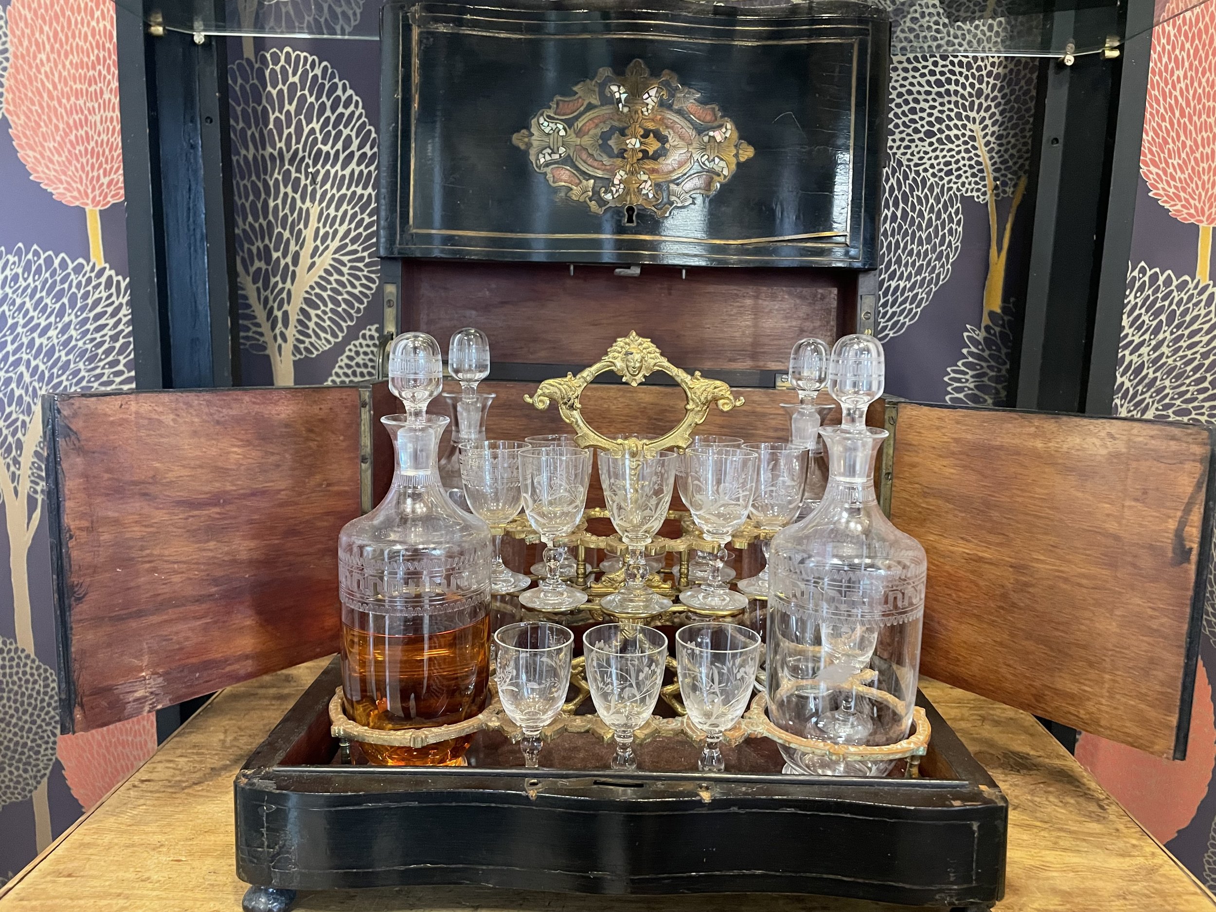 An ornate liquor decanter and glasses set on a wooden table with a vintage storage chest in the background.