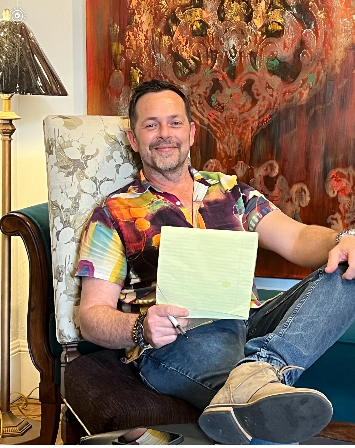 A man with short dark hair, beard, and light skin sitting in a patterned upholstered chair, holding a yellow notepad and pen, smiling at the camera. He is wearing a colorful, abstract-patterned shirt, jeans, and tan shoes. Behind him is a vibrant, abstract painting and beside him a patterned lampshade.