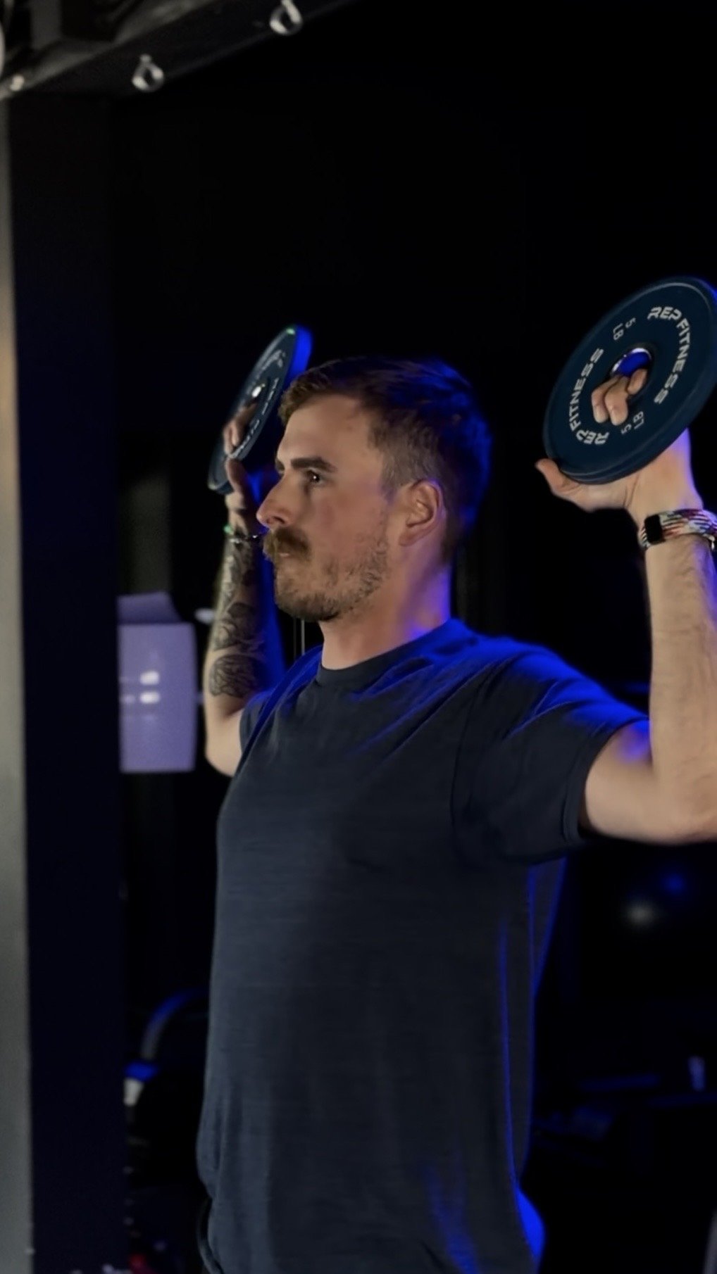 A man lifting black weight plates during a workout in a gym, wearing a dark t-shirt and wristwatch, with tattoos on his arm and a focused expression.