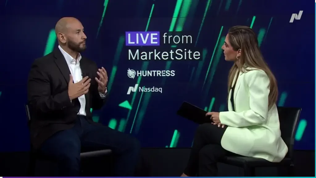 A man in a suit and a woman in a white blazer are seated and engaged in conversation on a television set. Behind them is a screen displaying the text "LIVE from MarketSite" and logos for Huntress and Nasdaq.