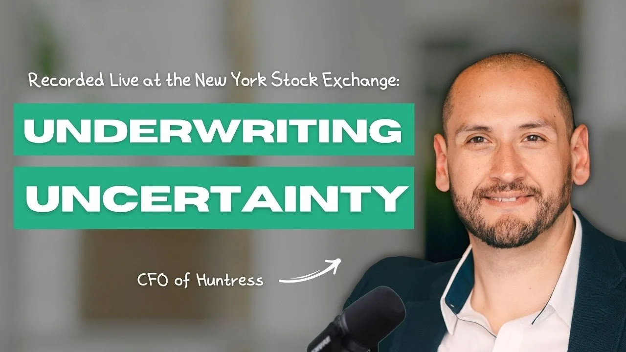 A man with a beard and a suit speaking at a microphone with a background of an office. Overlaid text reads: 'Recorded Live at the New York Stock Exchange: UNDERWRITING UNCERTAINTY'.