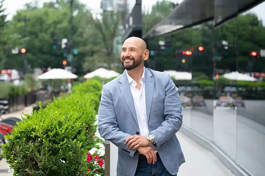Marcos Torres, leadership advisor and keynote speaker, standing outdoors near a glass building with greenery and outdoor seating in the background.