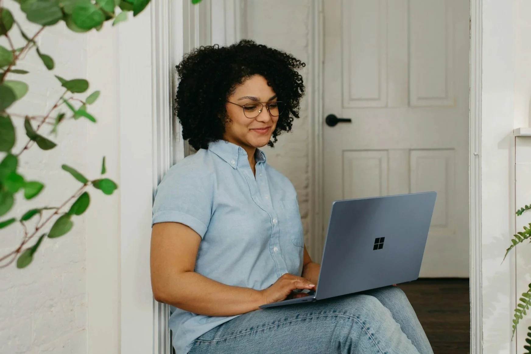 A woman sitting on the floor near a doorway, working on a silver Microsoft Surface laptop, surrounded by green plants, indoors.