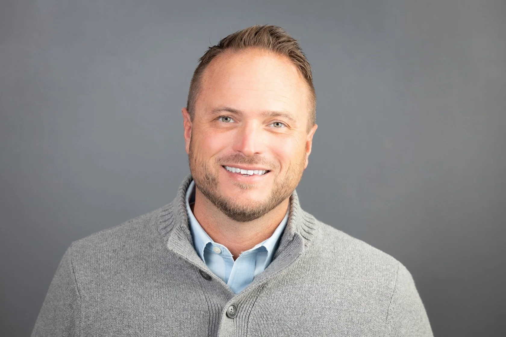 A smiling man with light skin, short brown hair, light-colored eyes, light beard, wearing a light blue shirt and gray sweater, against a gray background.