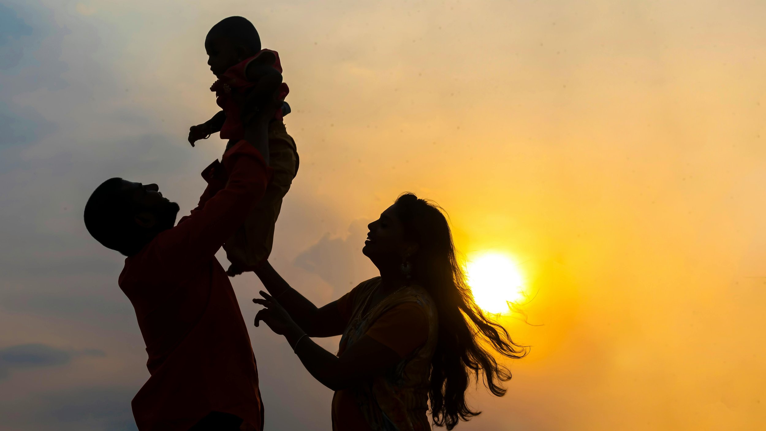 Silhouettes of a family playing together during sunset, with a man and woman lifting a child in the air against a yellow-orange sky.