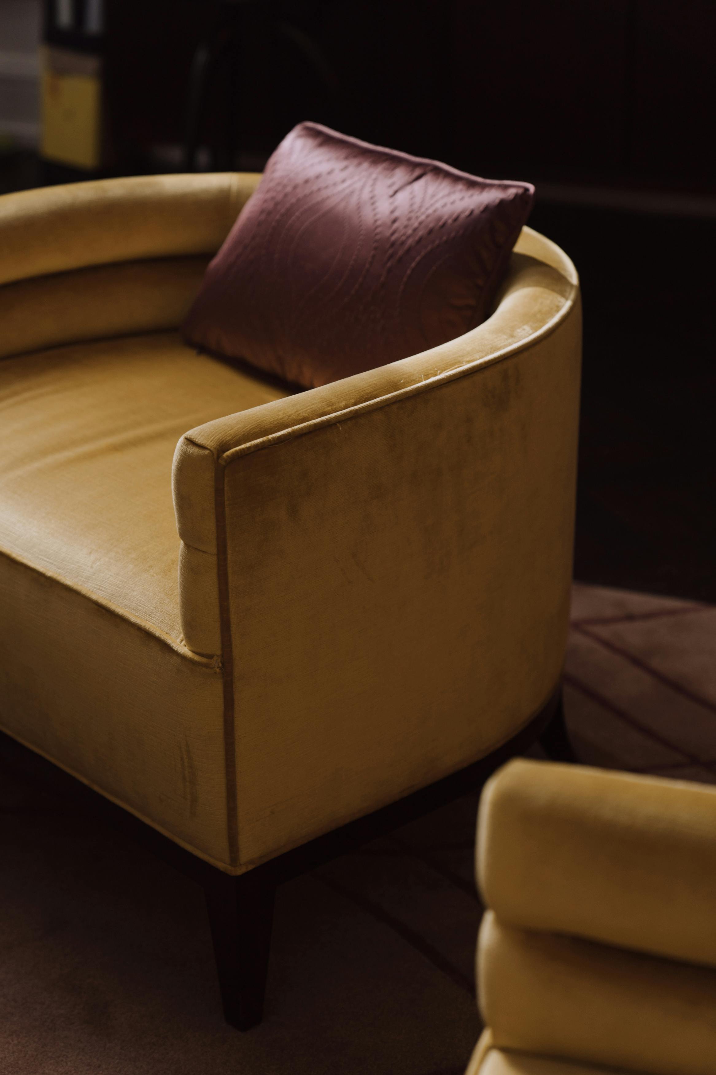 A mustard yellow velvet armchair with wooden legs and a maroon pillow resting against the back, in a dimly lit room.