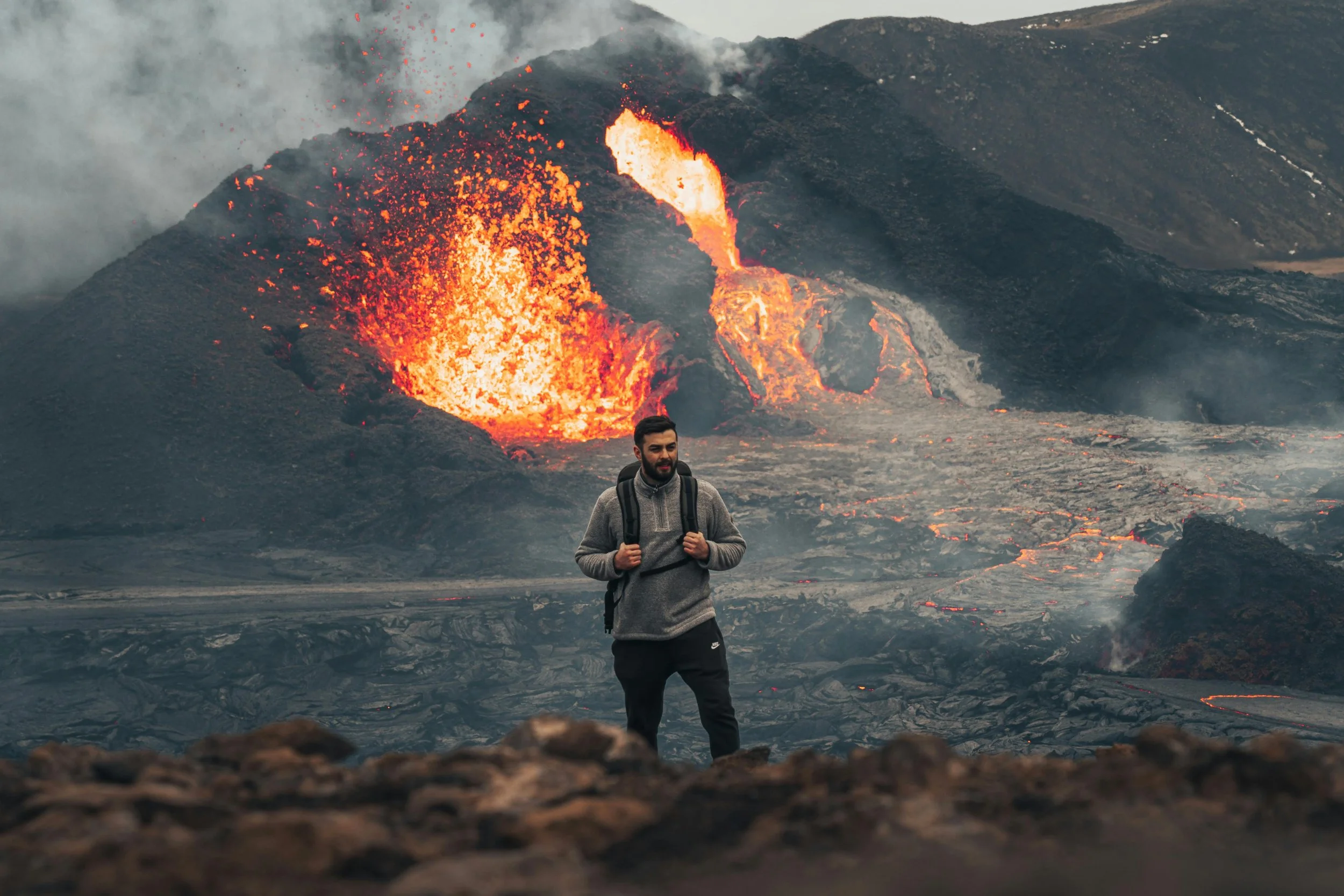A man with a backpack standing on rocky terrain with erupting volcano and flowing lava in the background.