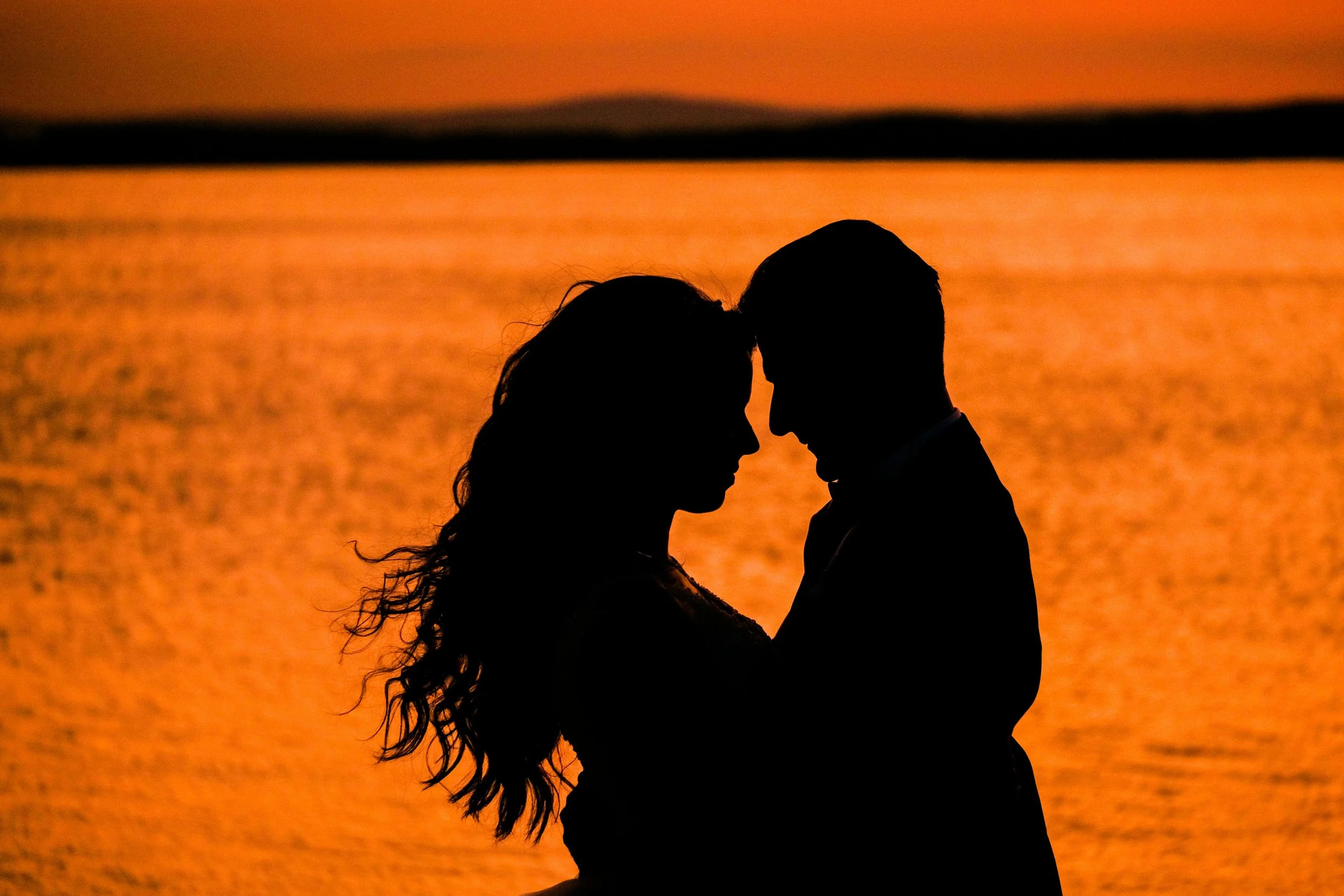 Silhouetted couple embracing by a sunset over water, with the sun low on the horizon and colorful sky
